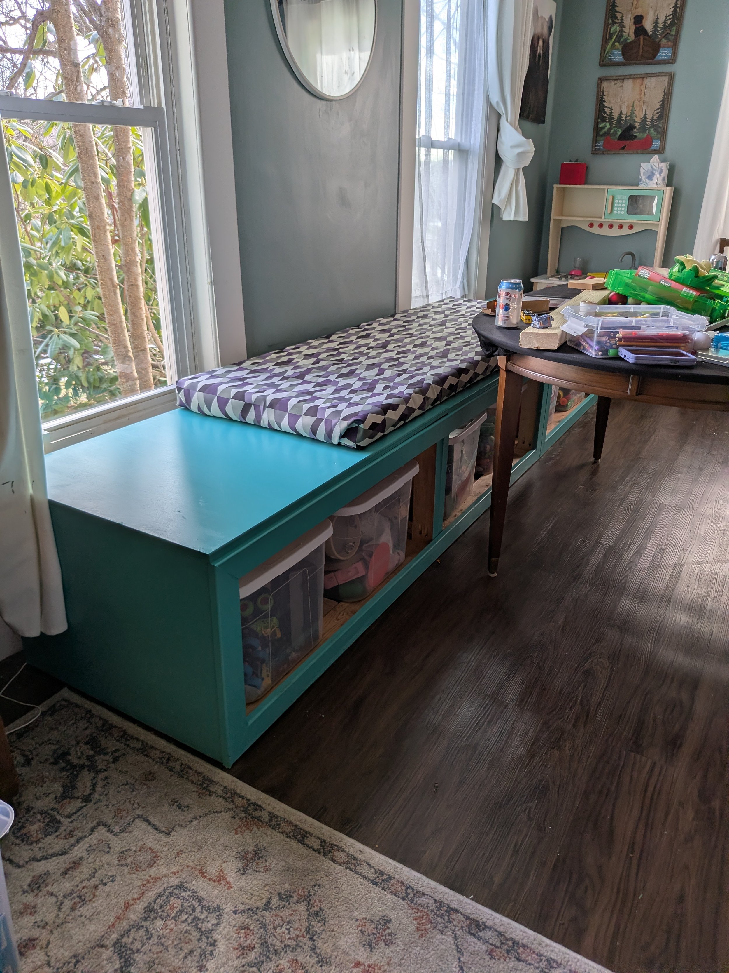 A turquoise window seat with a geometric patterned cushion, beneath a large window with white curtains, in a room decorated with wall art and toys, with a wooden table holding various items and a floor with a patterned rug and dark wood flooring.