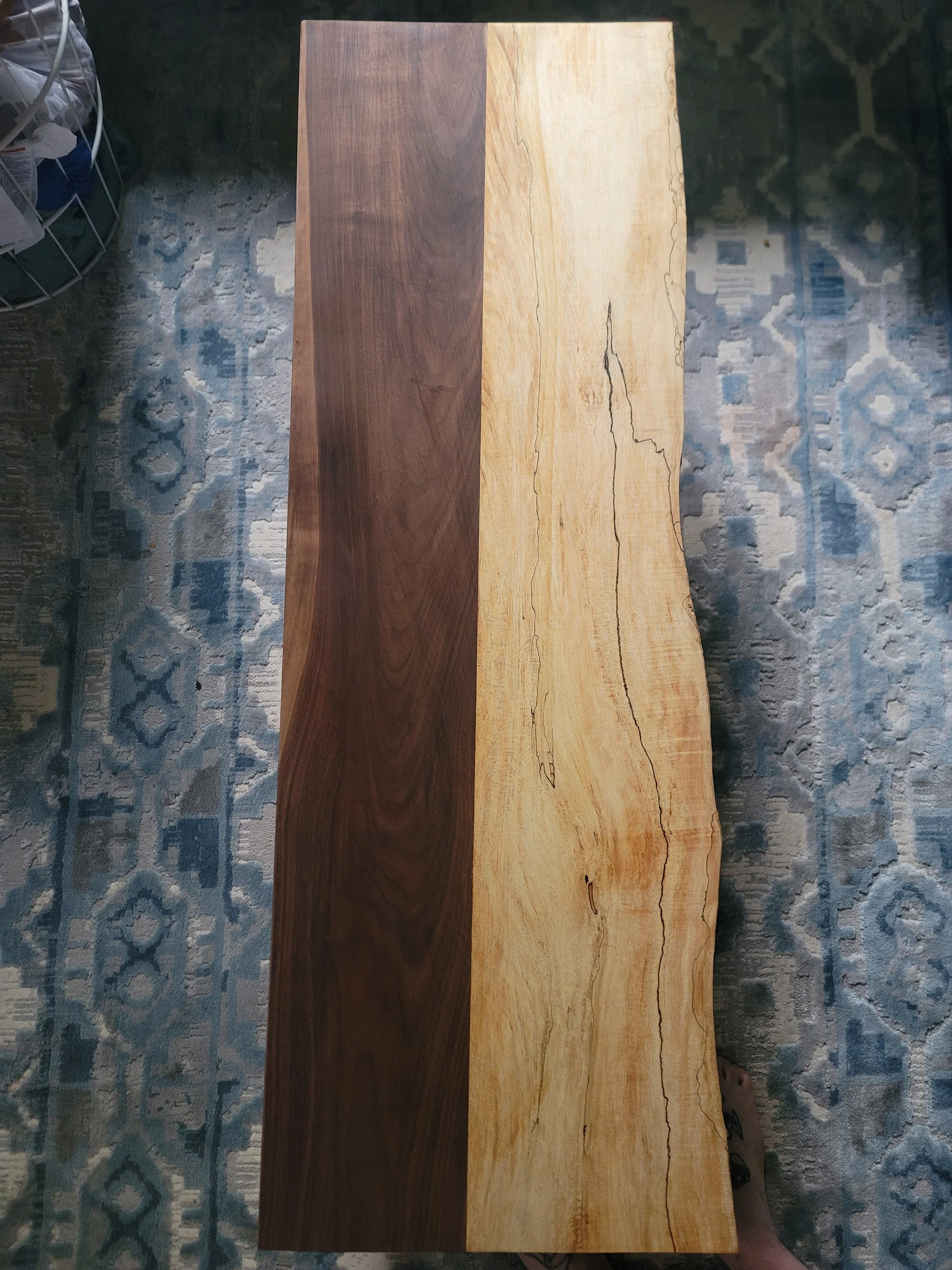 A wooden table with a natural, live edge, showing two different types of wood with contrasting colors and grain patterns, placed on a patterned blue and gray rug.