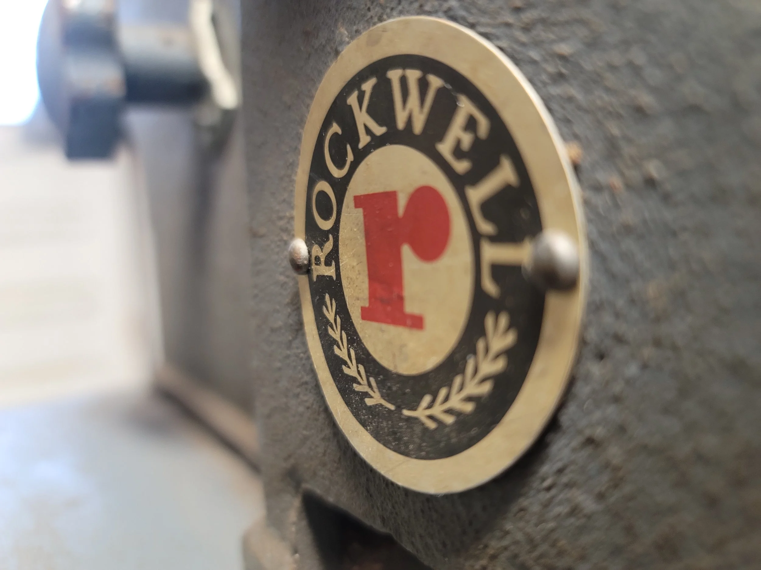 Close-up of a circular brass badge with black and red markings reading 'Rockwell 1' and small decorative leaf accents.
