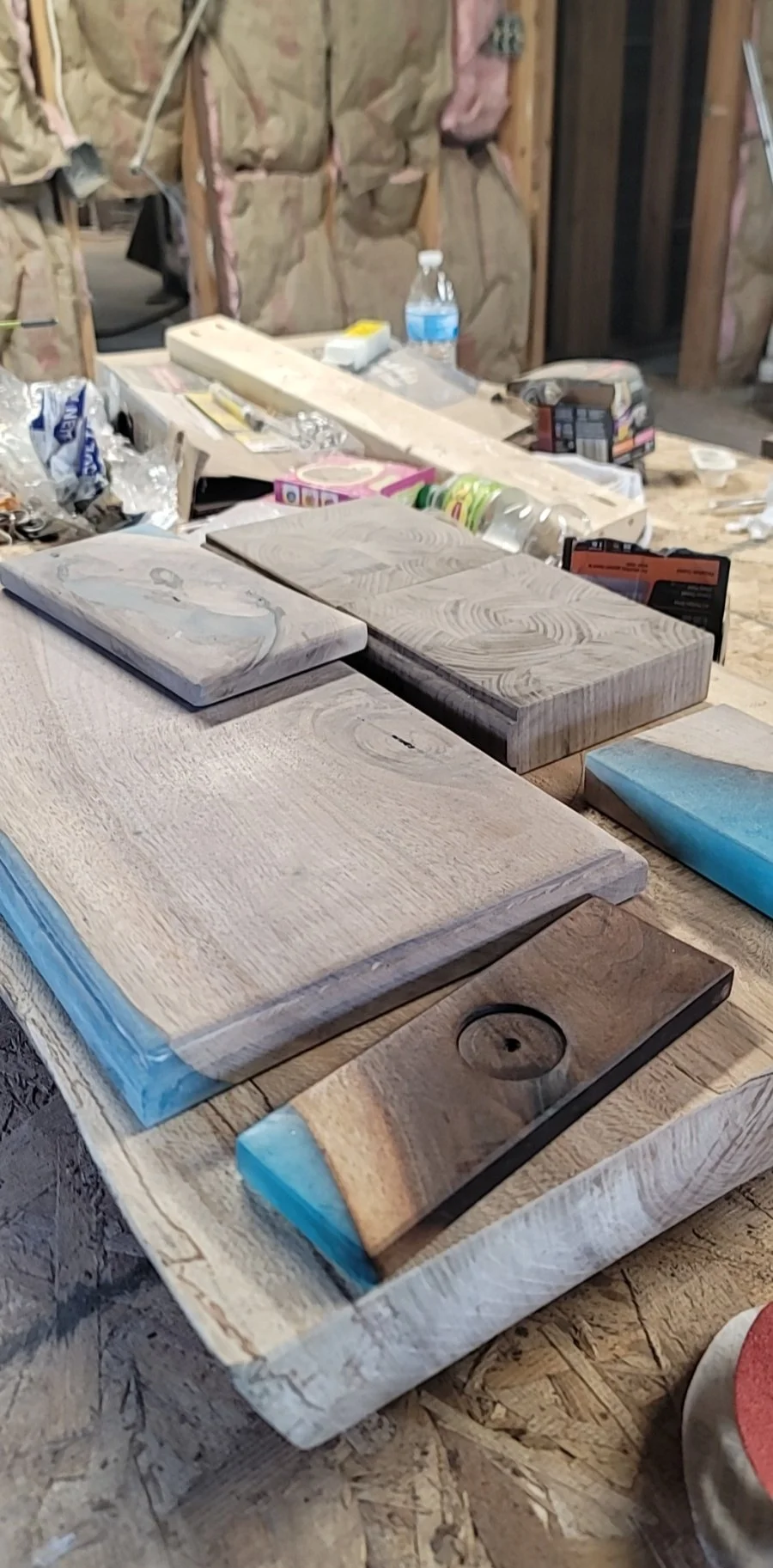 A woodworking workspace with pieces of wood and epoxy inlay on a workbench, surrounded by tools, water bottles, and various workshop supplies.