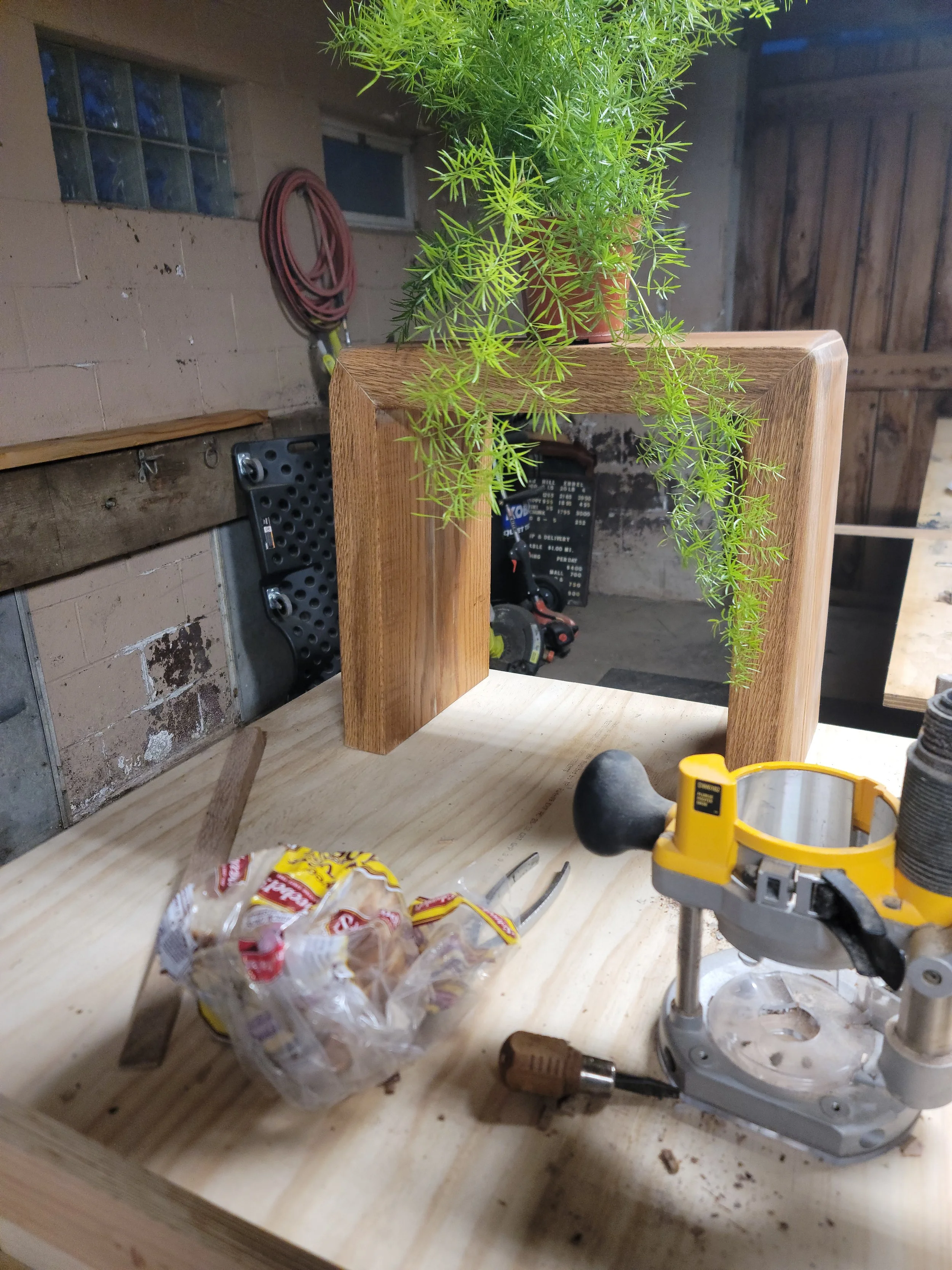 A woodworking workshop with a wooden frame supporting a potted plant, a bag of snacks, hand tools, and a yellow router on a wooden workbench.
