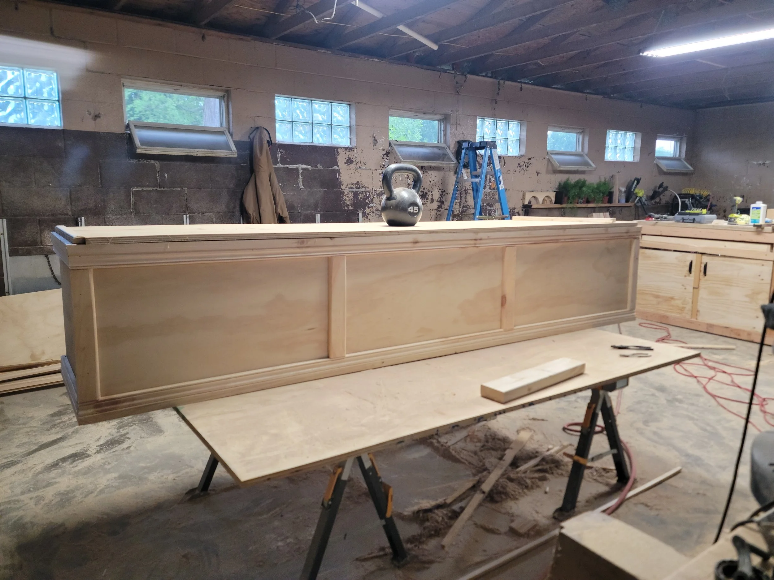 A woodworking workshop with a long wooden cabinet in progress placed on a work table, with a kettlebell on top, surrounded by tools and materials.