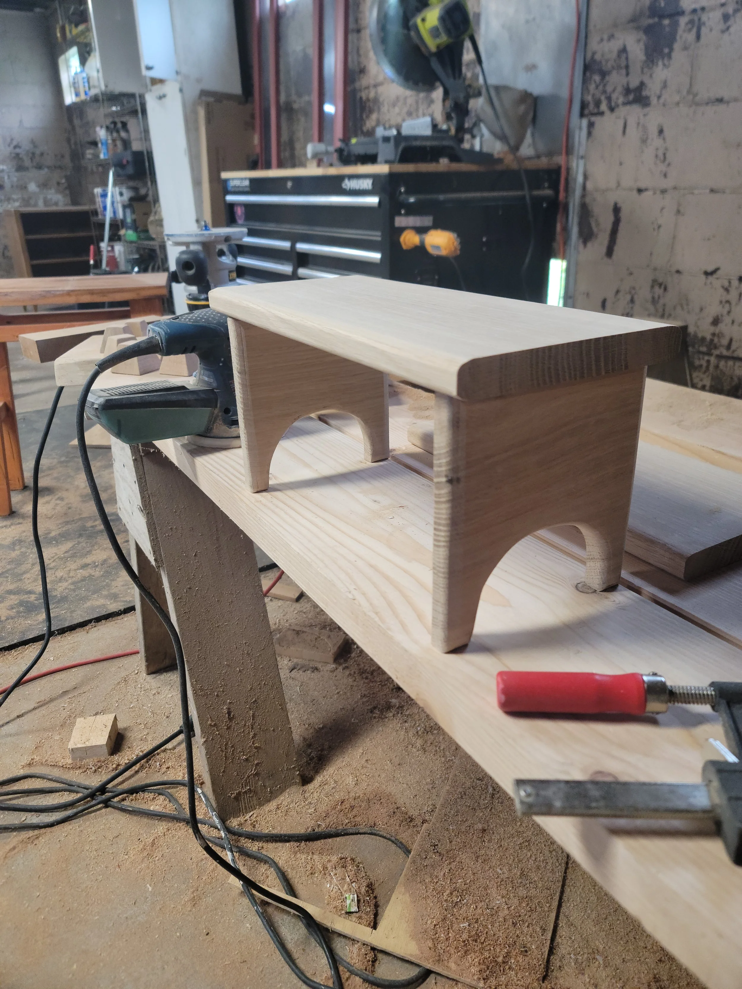 A small wooden bench or stool in progress on a workbench in a woodworking shop.
