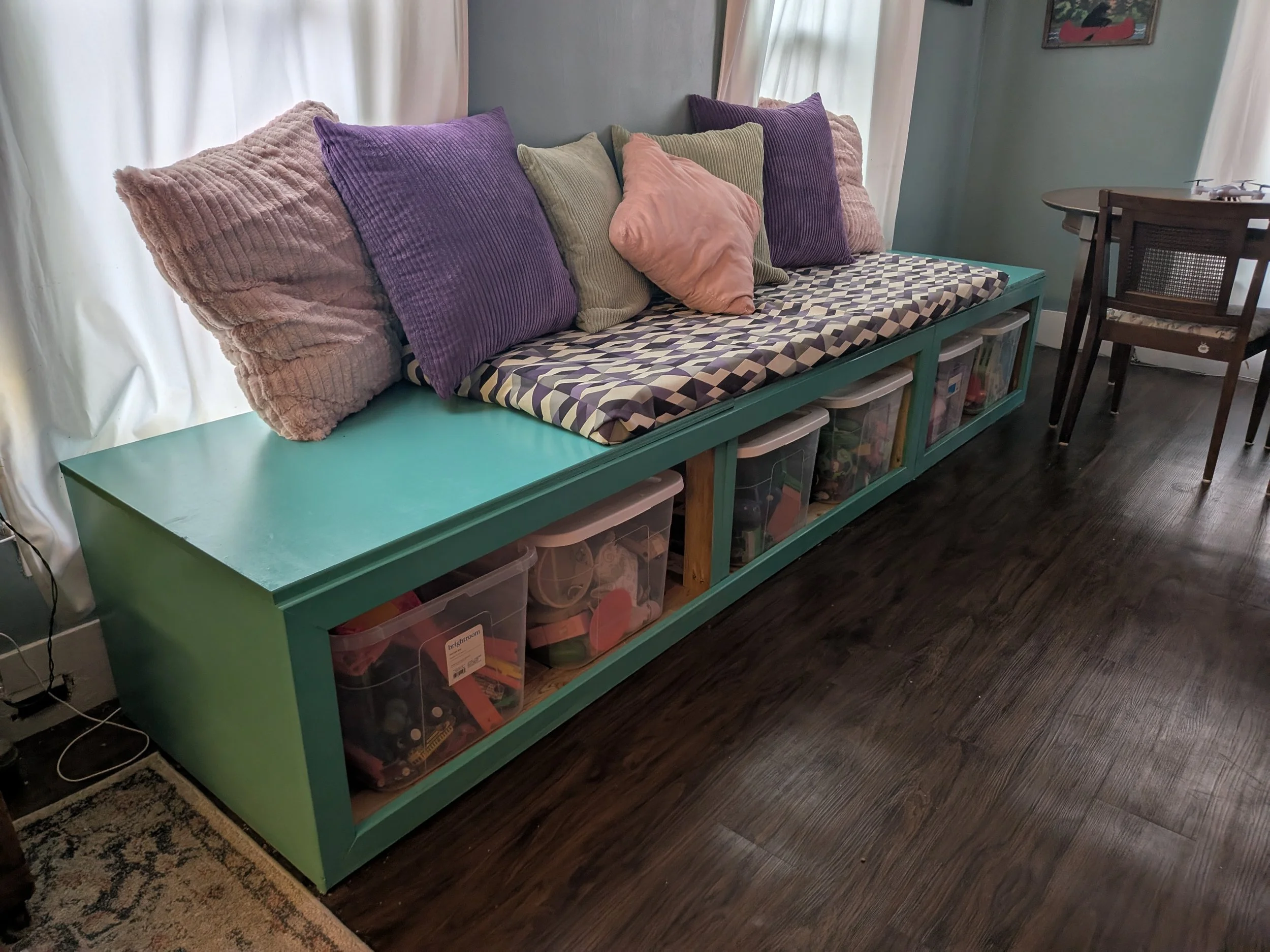 A teal-colored storage bench with a geometric-patterned cushion and multiple purple, green, and pink pillows on top. The bench contains clear plastic bins filled with toys, sitting next to a dark wooden dining table and chairs in a room with dark woo
