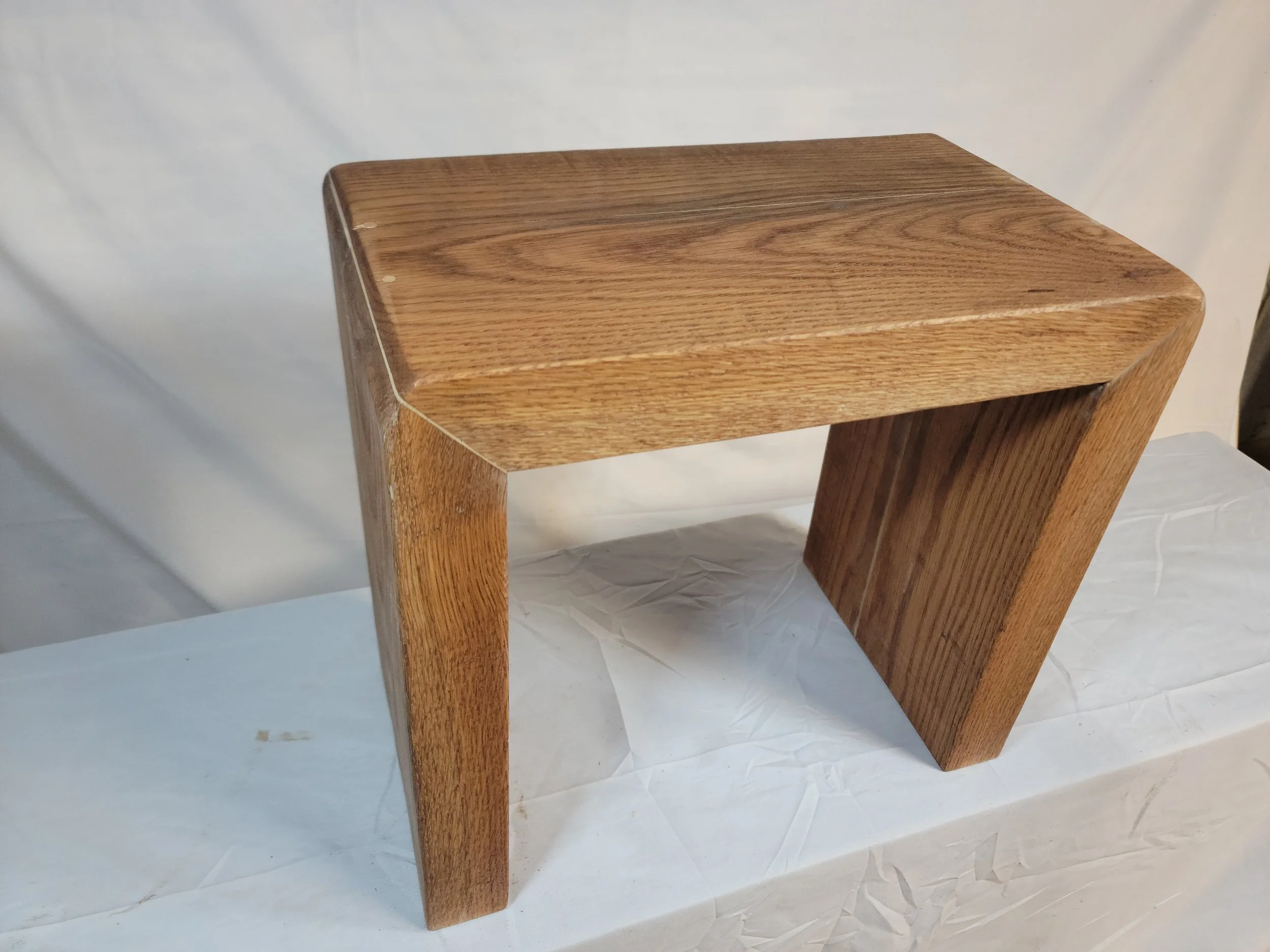 A small wooden bench or stool with a rectangular seat and two supporting legs on each side, finished with a natural wood stain.