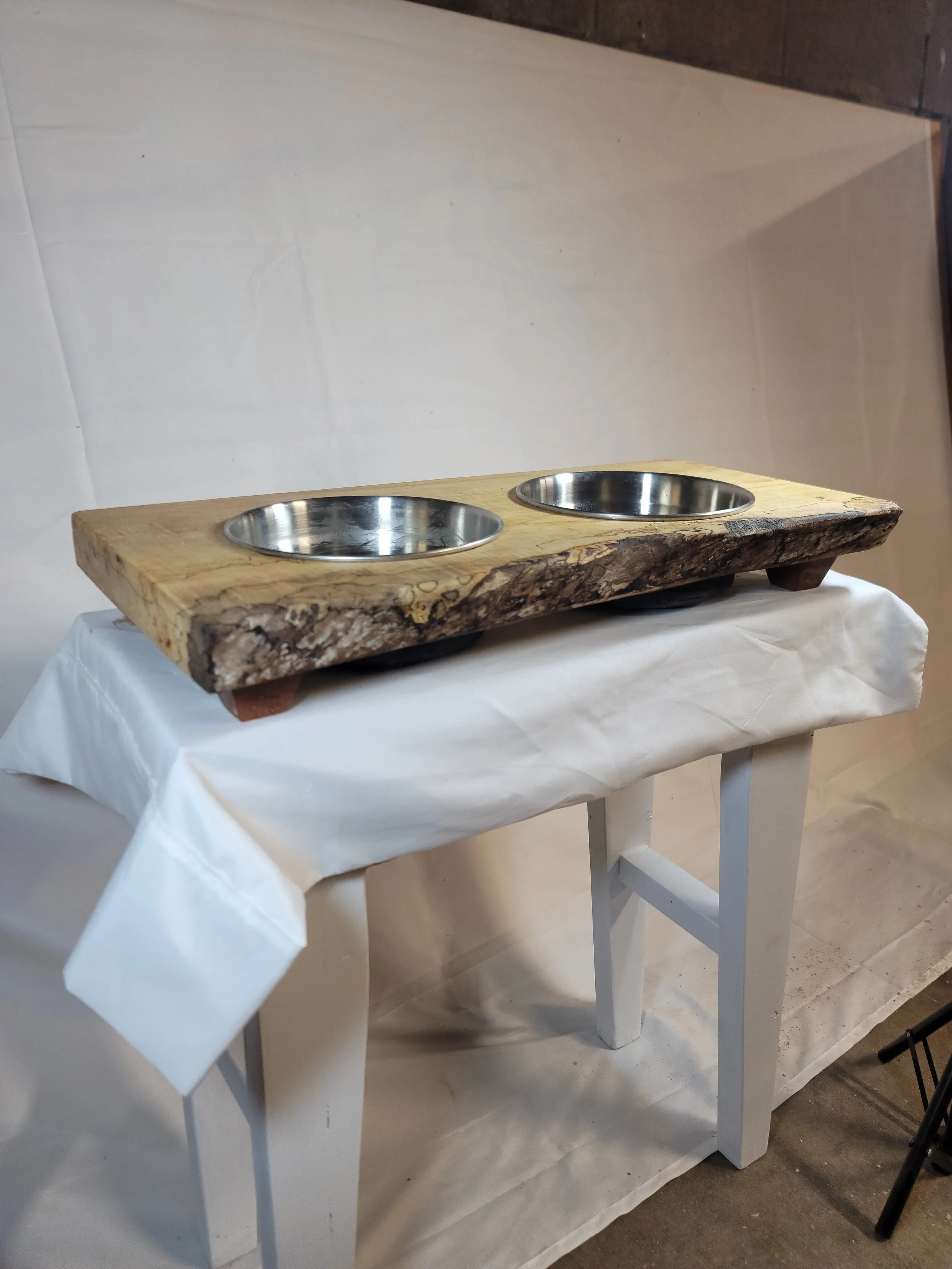 A rustic wooden pet feeding station with two round stainless steel bowls, placed on a white table covered with white paper or cloth, against a plain background.