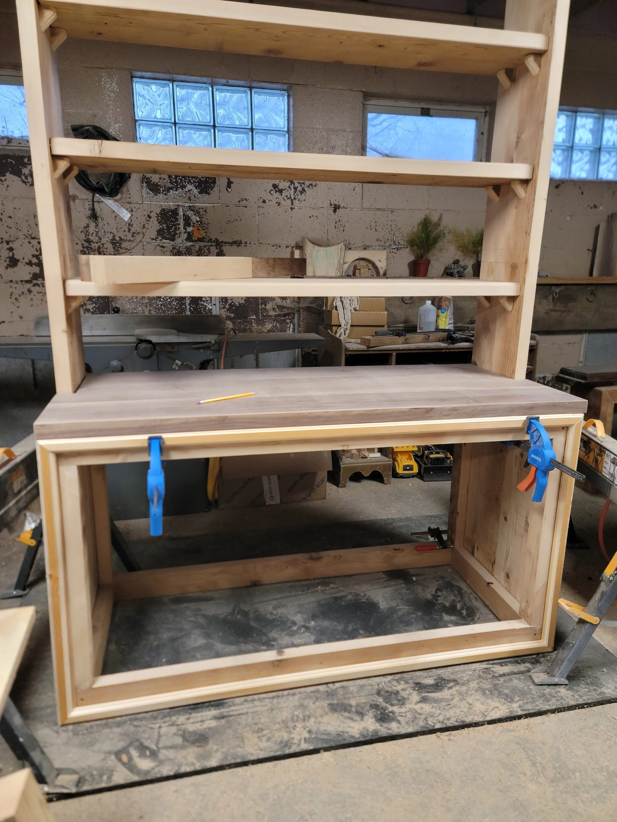 A woodworking project in progress featuring a partially assembled wooden workbench with a shelving unit on top, clamps on the sides, and a pencil on the work surface in a workshop setting.