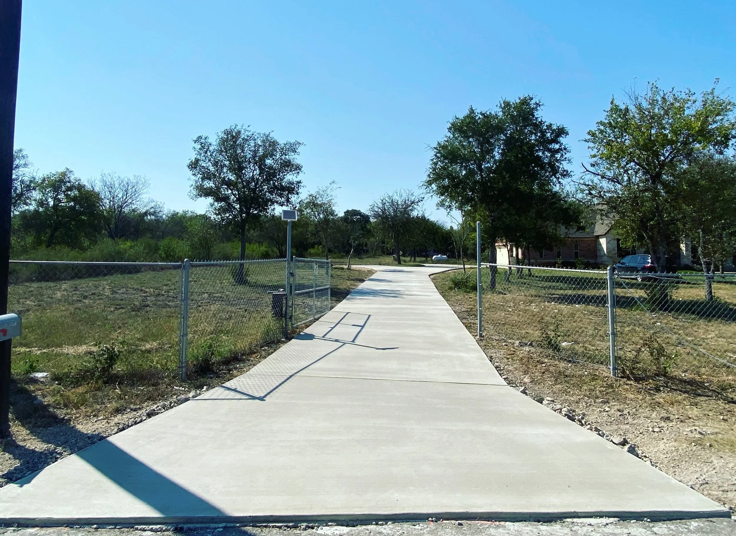 Newly poured concrete sidewalk in a residential neighborhood with trees, houses, a fence, and a car in the background.
