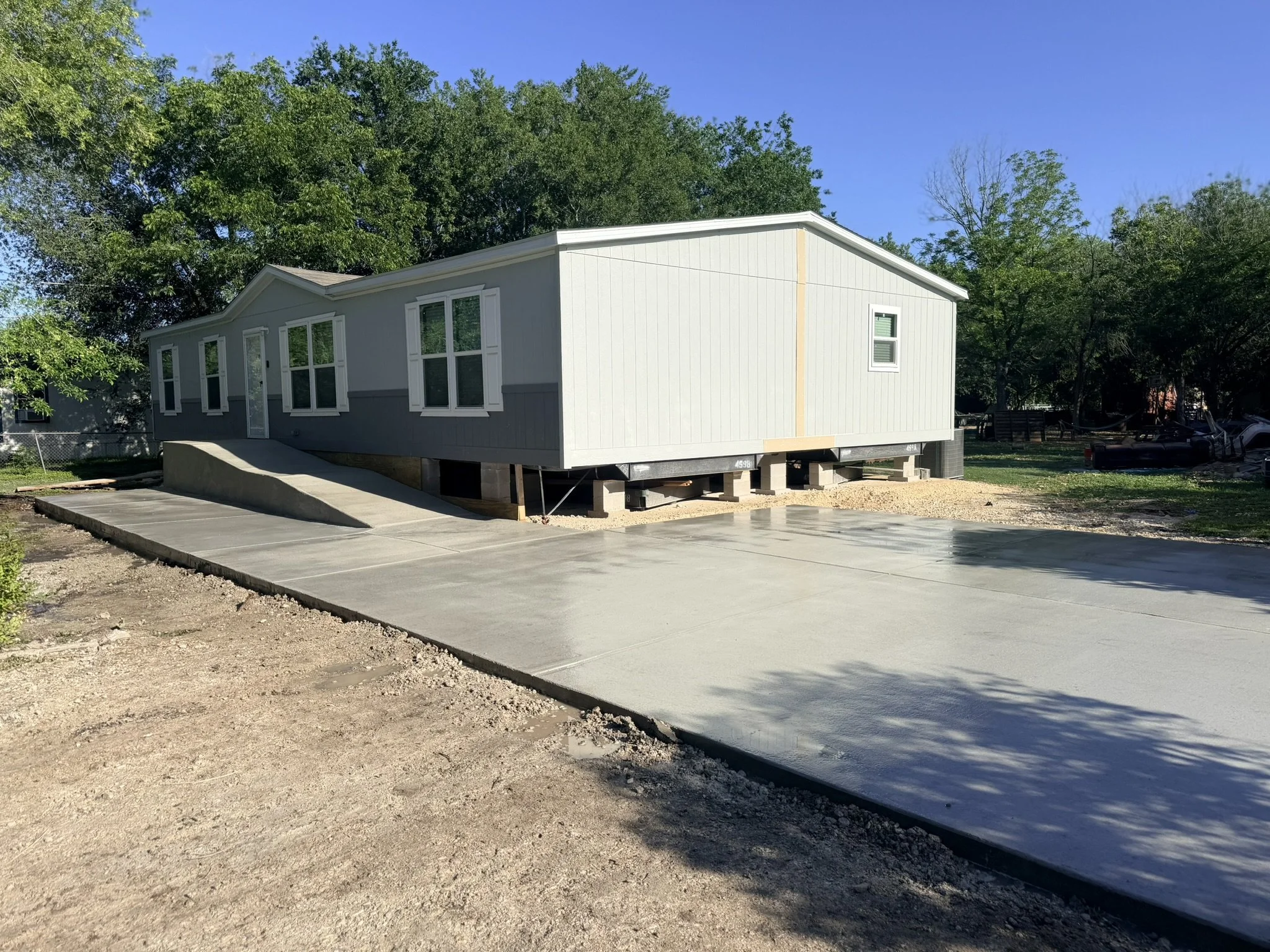 A house under construction with a concrete foundation and a wood frame, surrounded by trees and a partly cloudy sky.