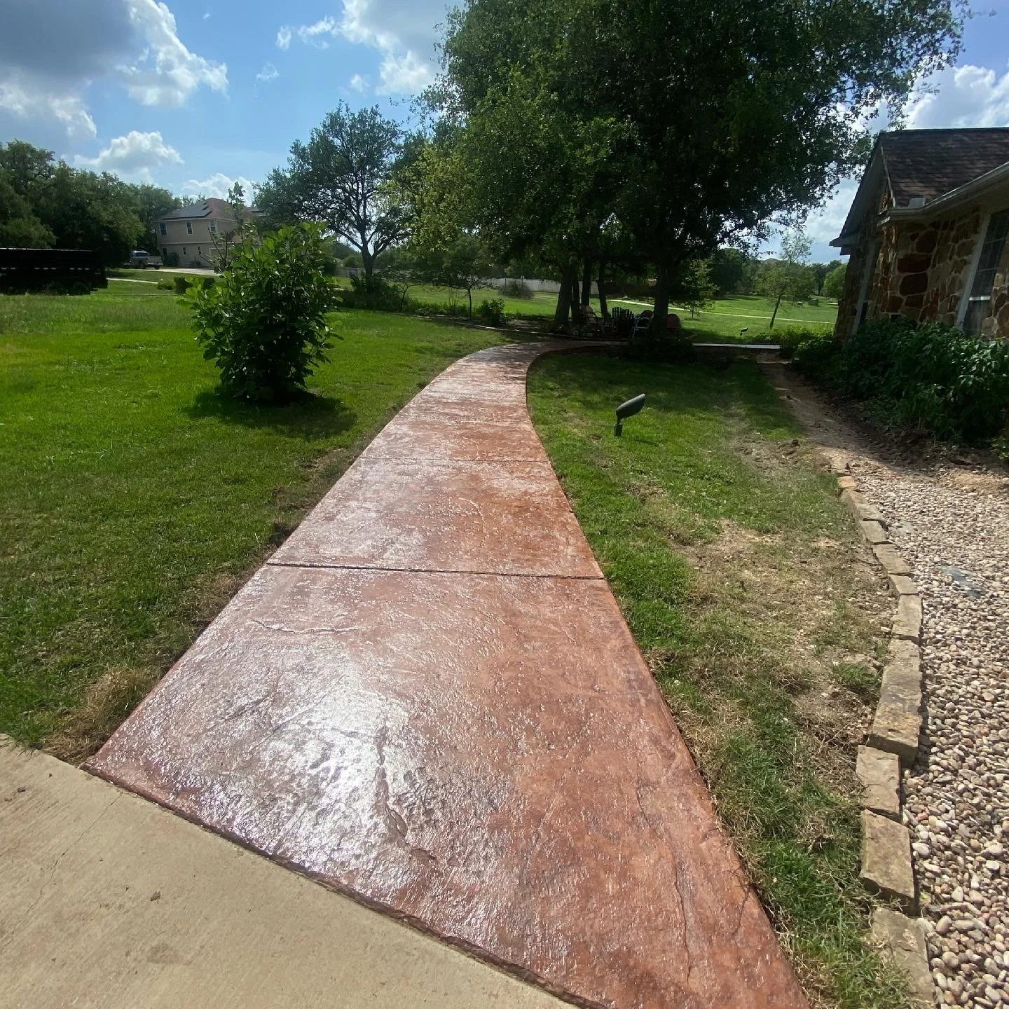 A curved, freshly painted red concrete sidewalk in a backyard, with a grassy lawn on both sides, trees in the background, and a house with stone walls on the right.