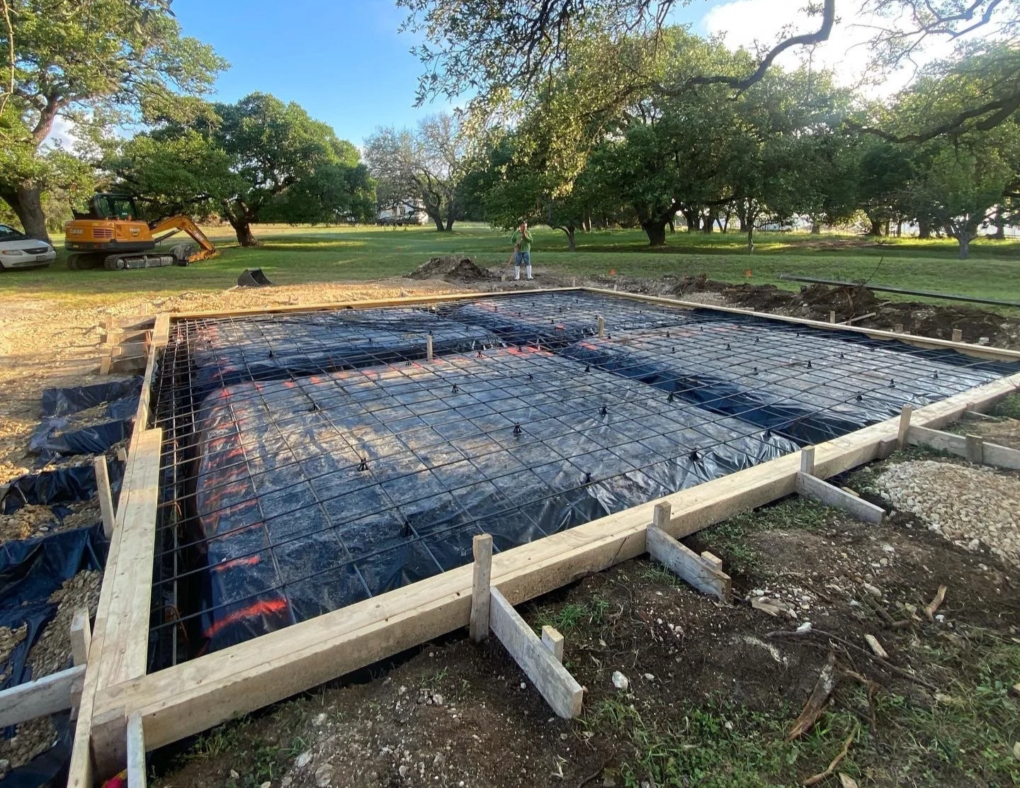 Construction site with wooden frame and metal rebar for a concrete slab, black plastic sheeting underneath, in a grassy park with trees and a small excavator in the background.
