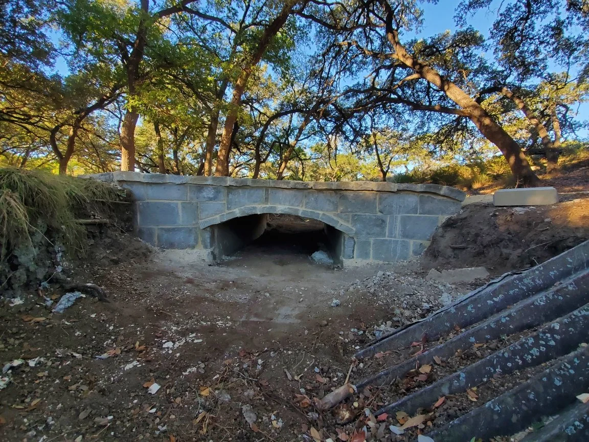 Under-construction stone bridge in a wooded area with trees and dirt road.