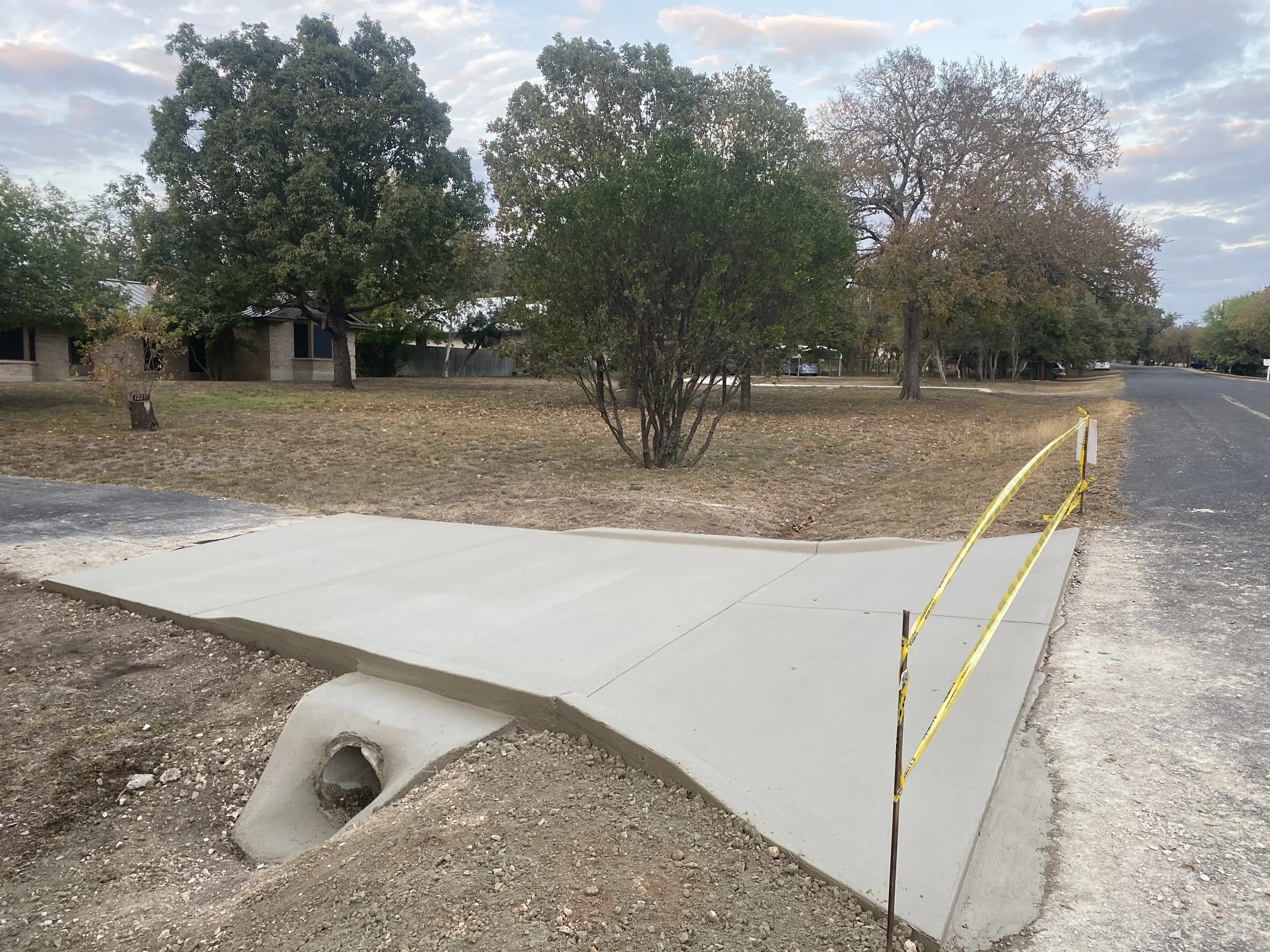 A newly poured concrete sidewalk section under construction, surrounded by dirt and construction barriers in a residential area with green trees and houses in the background.