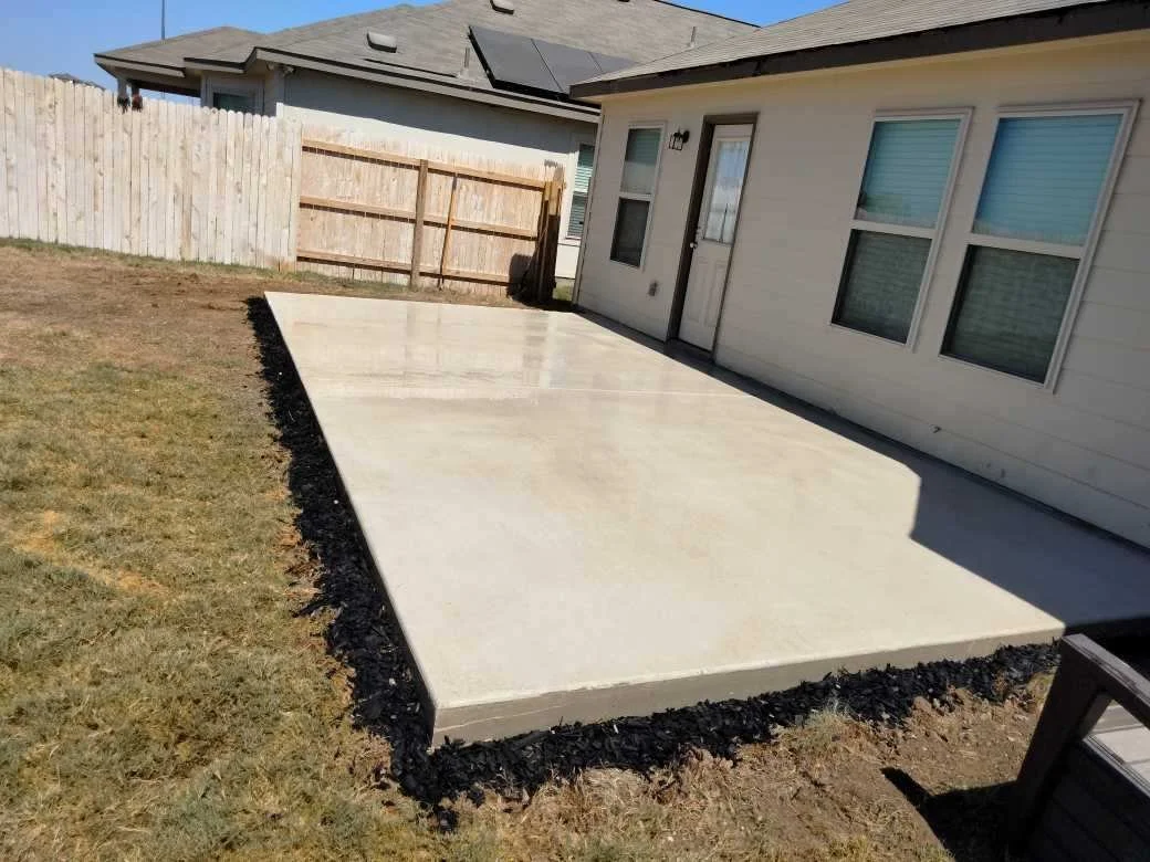 Newly poured concrete patio in backyard next to house with white siding, three windows, and a door, surrounded by black edging and a patch of grass, enclosed by a wooden fence.