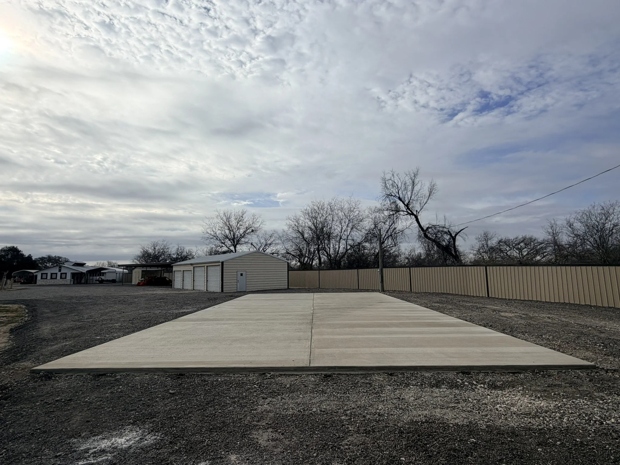 A newly poured concrete sidewalk in a residential area with older trees, fences, and houses, under a cloudy sky.