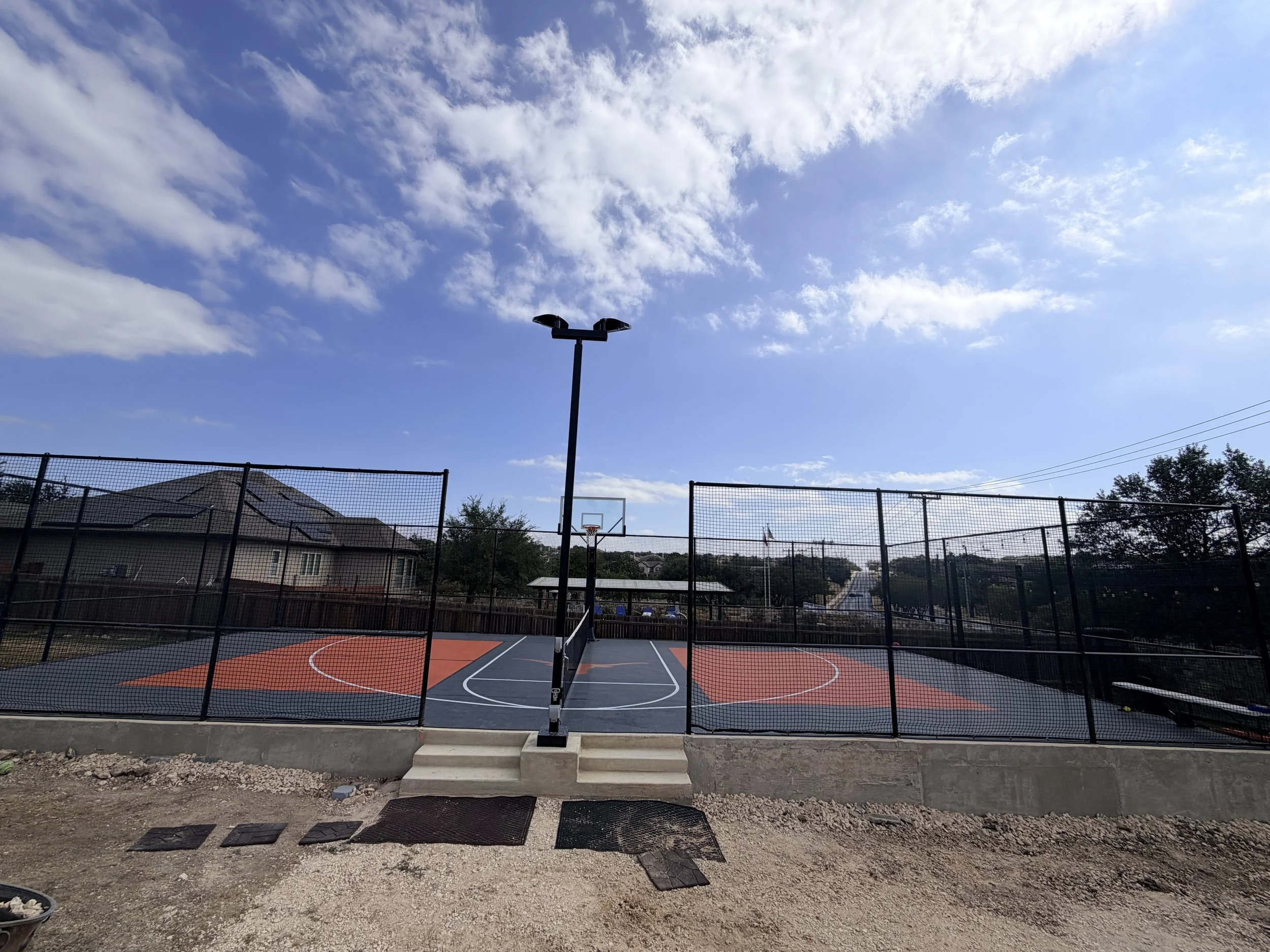 An outdoor basketball court with a fence, basketball hoops, and a black light pole underneath a partly cloudy blue sky with white clouds.