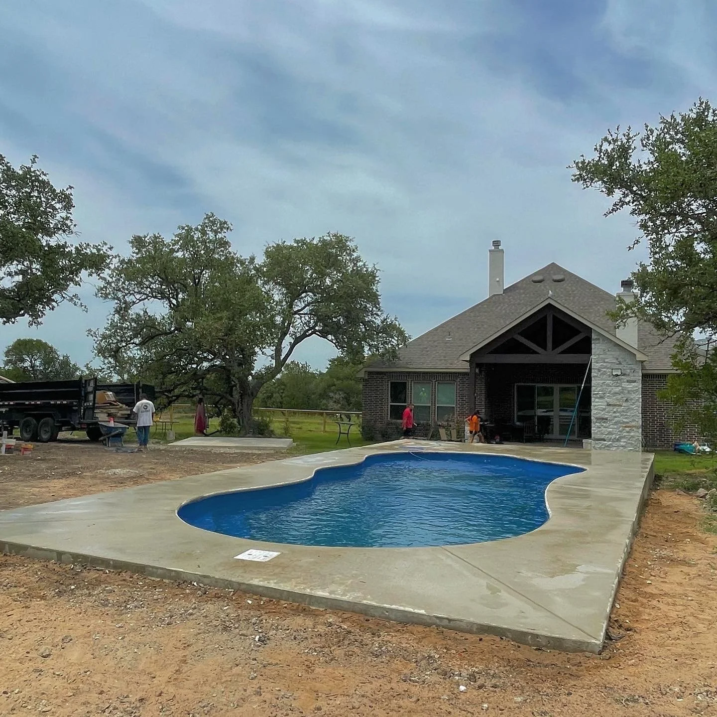 Construction site of a backyard swimming pool with a new concrete deck, a house with a screened porch, and workers installing the pool in a suburban backyard with trees and a cloudy sky.