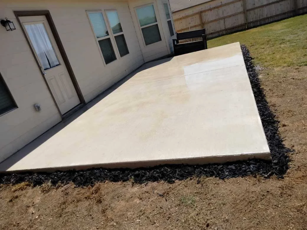 Backyard patio made of concrete with a black mulch border, adjacent to a house with a door and several windows, and a grassy area with a wooden fence in the background.
