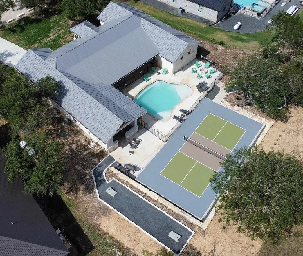 Aerial view of a house with a gray metal roof, a swimming pool, a tennis court, and outdoor seating areas.