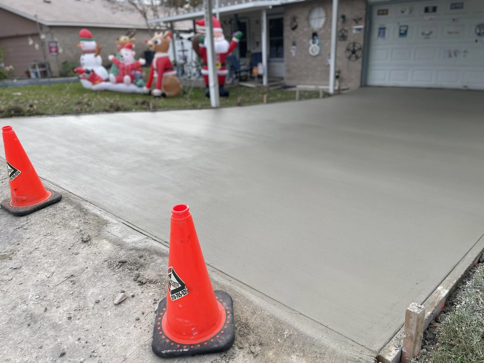 Orange construction cones on a newly poured concrete driveway in front of a house decorated for Christmas with inflatable reindeer, snowmen, and Santa Claus, and various holiday decorations on the house.