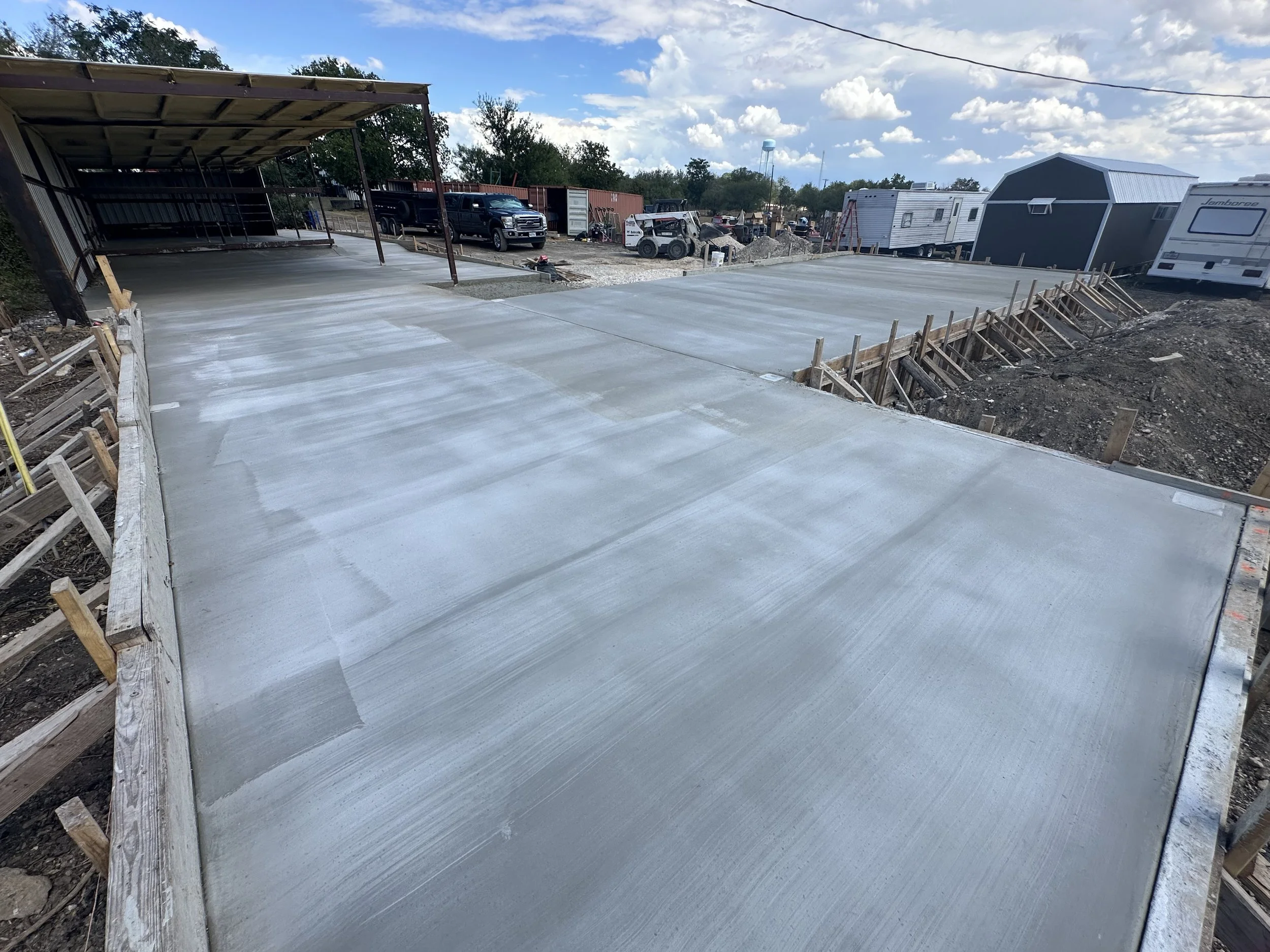 Newly poured concrete driveway with framing and formwork in place, surrounded by construction materials and vehicles, with RVs and trees in the background under a partly cloudy sky.