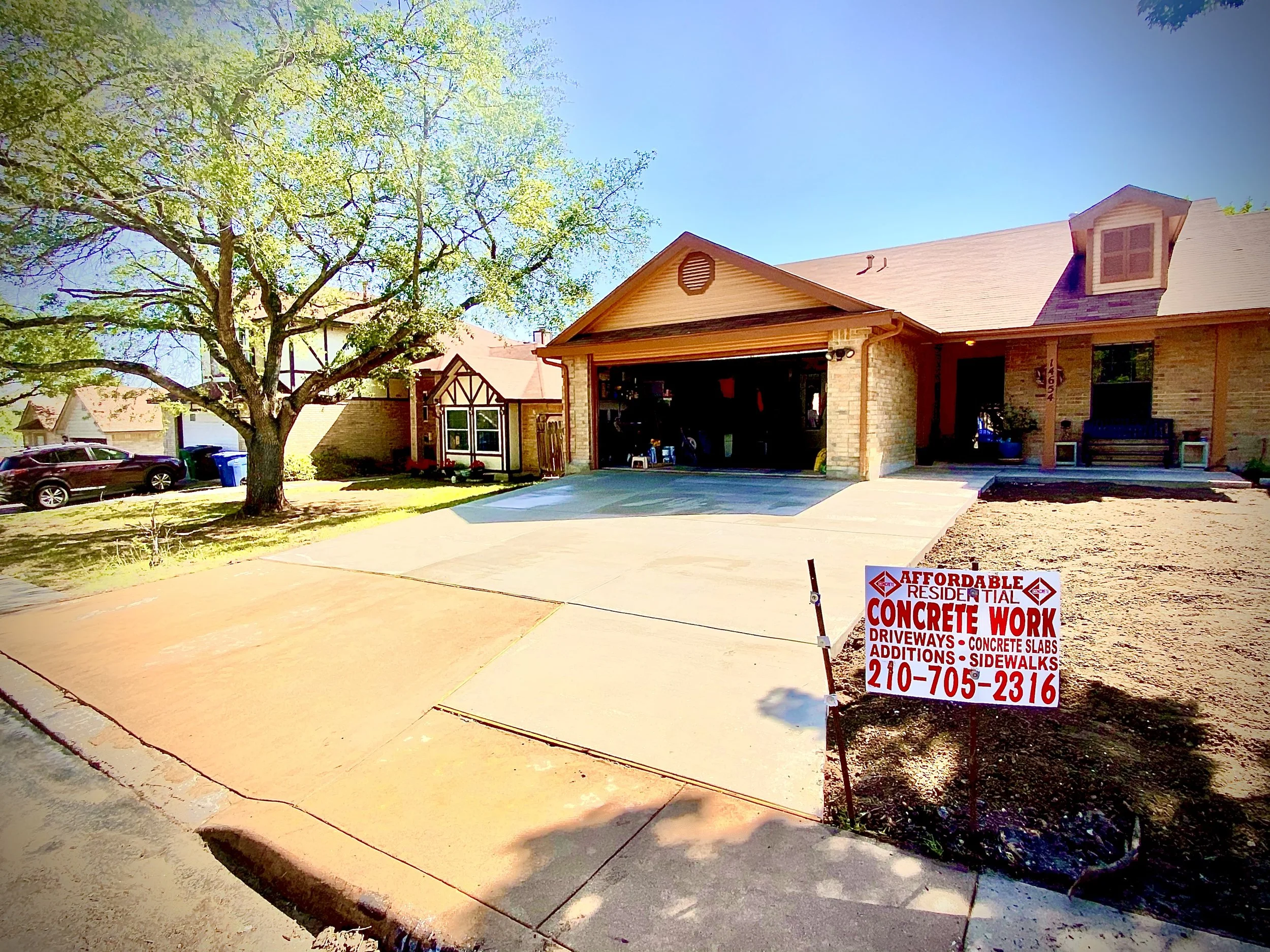 A residential house undergoing concrete work with a sign advertising concrete services, parked cars, a tree, and a clear blue sky.