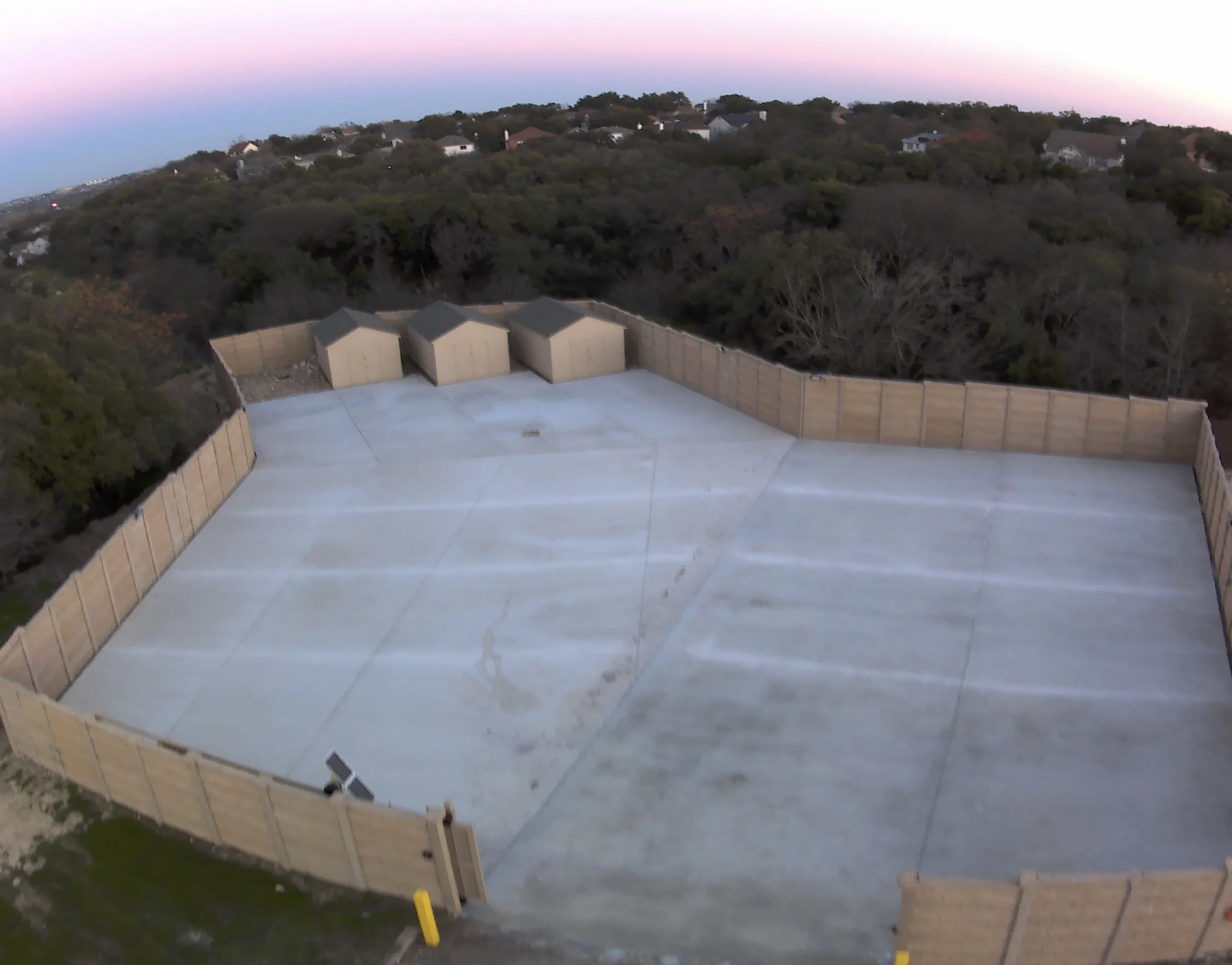 View from a rooftop parking lot with four small wooden storage sheds, beige walls, and a surrounding wooden fence, overlooking a hillside residential area with houses and trees at dusk.