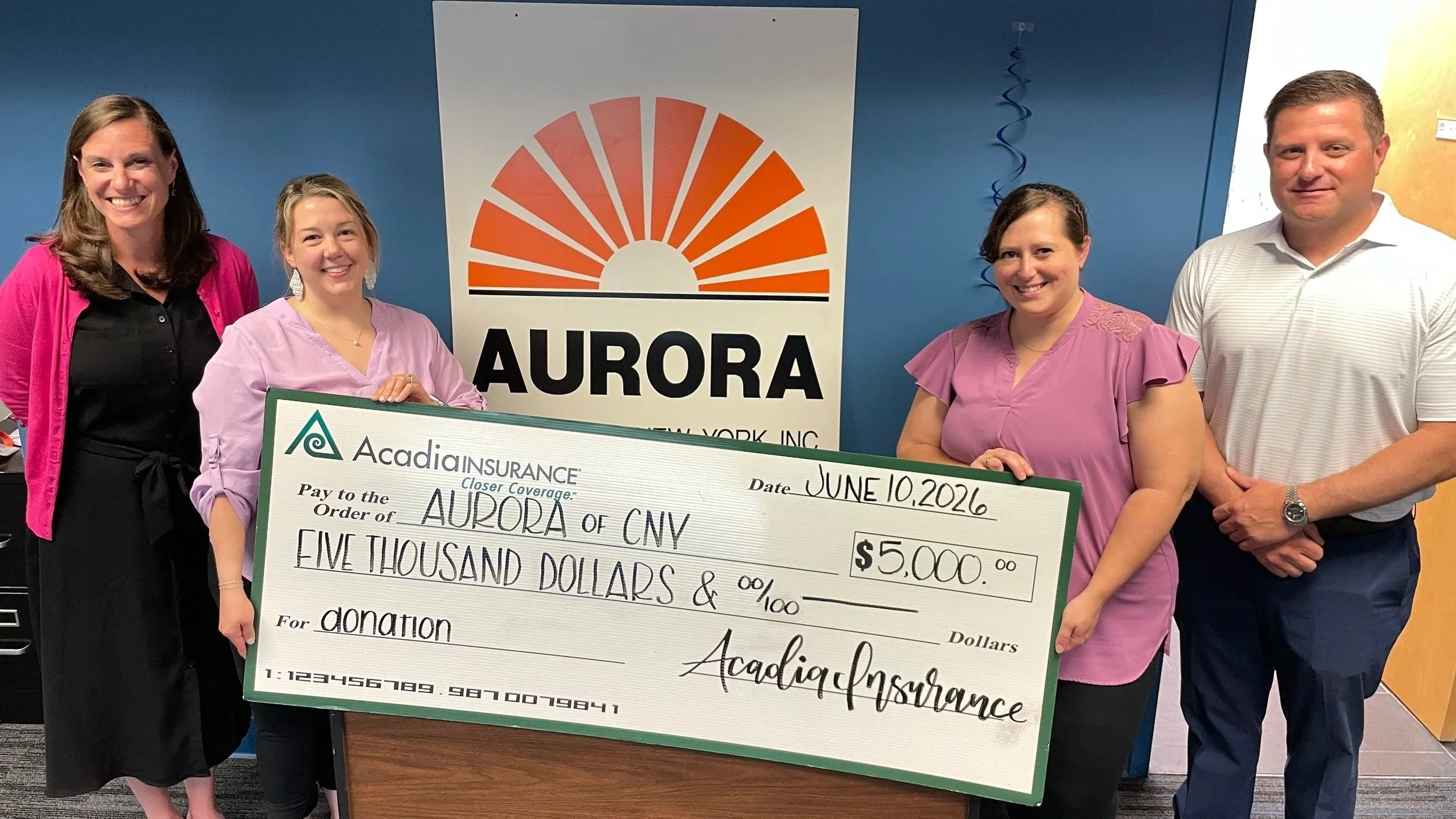 A group of four people pose with a large donation check from Acadia Insurance for $5,000. Two women are visible to the left of the check, a woman and a man are visible to the right.