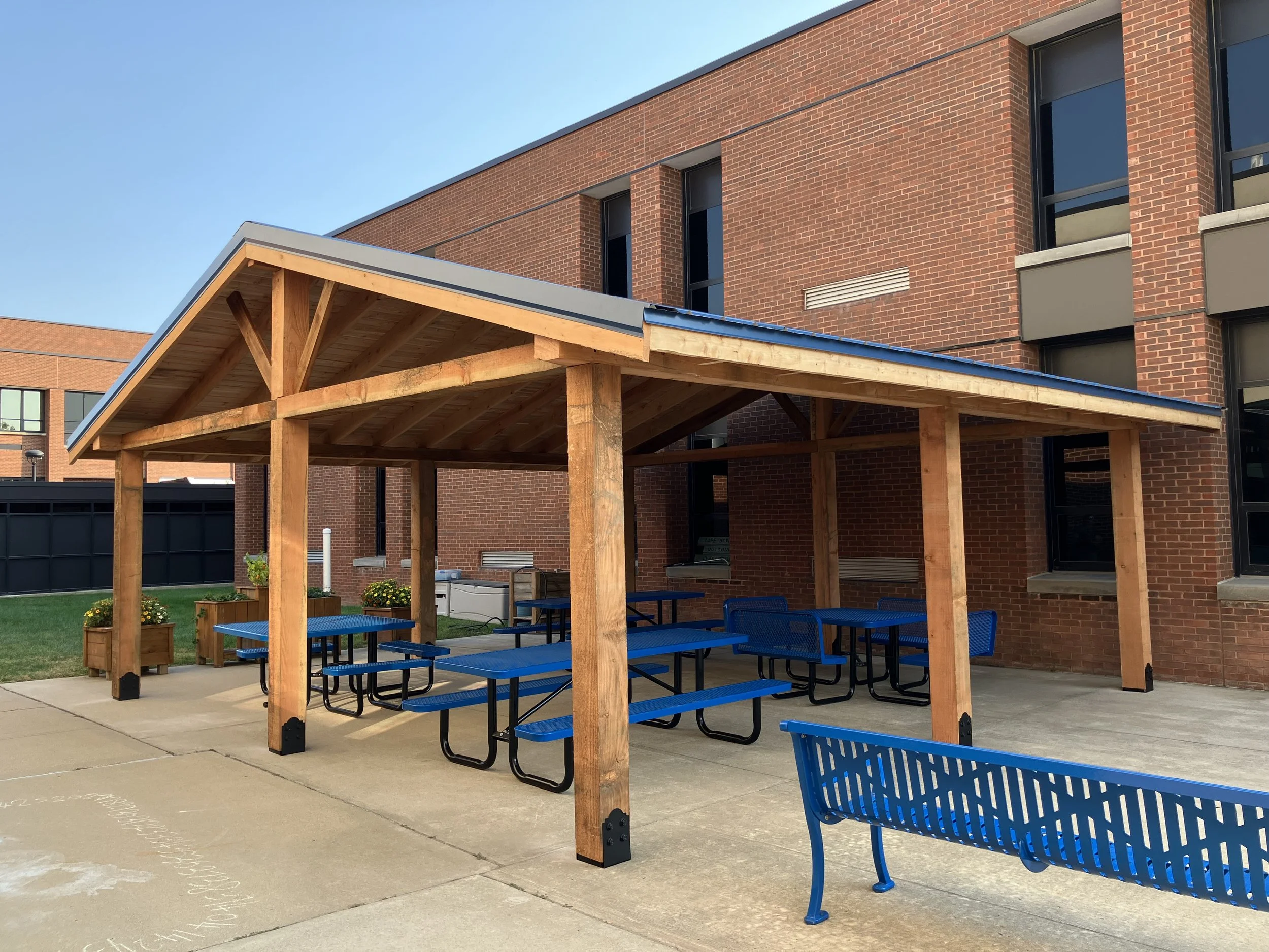 Outdoor patio area with wooden pergola, blue picnic tables and benches, a blue metal bench, potted plants, and a brick building in the background.