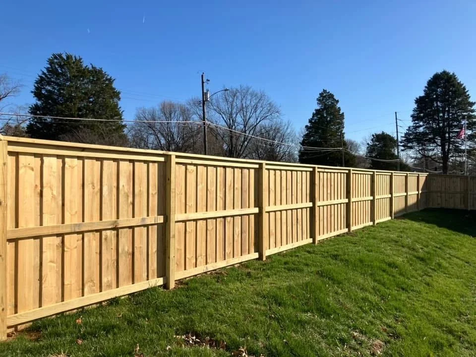 New wooden fence in a backyard with green grass, trees, and a clear blue sky.