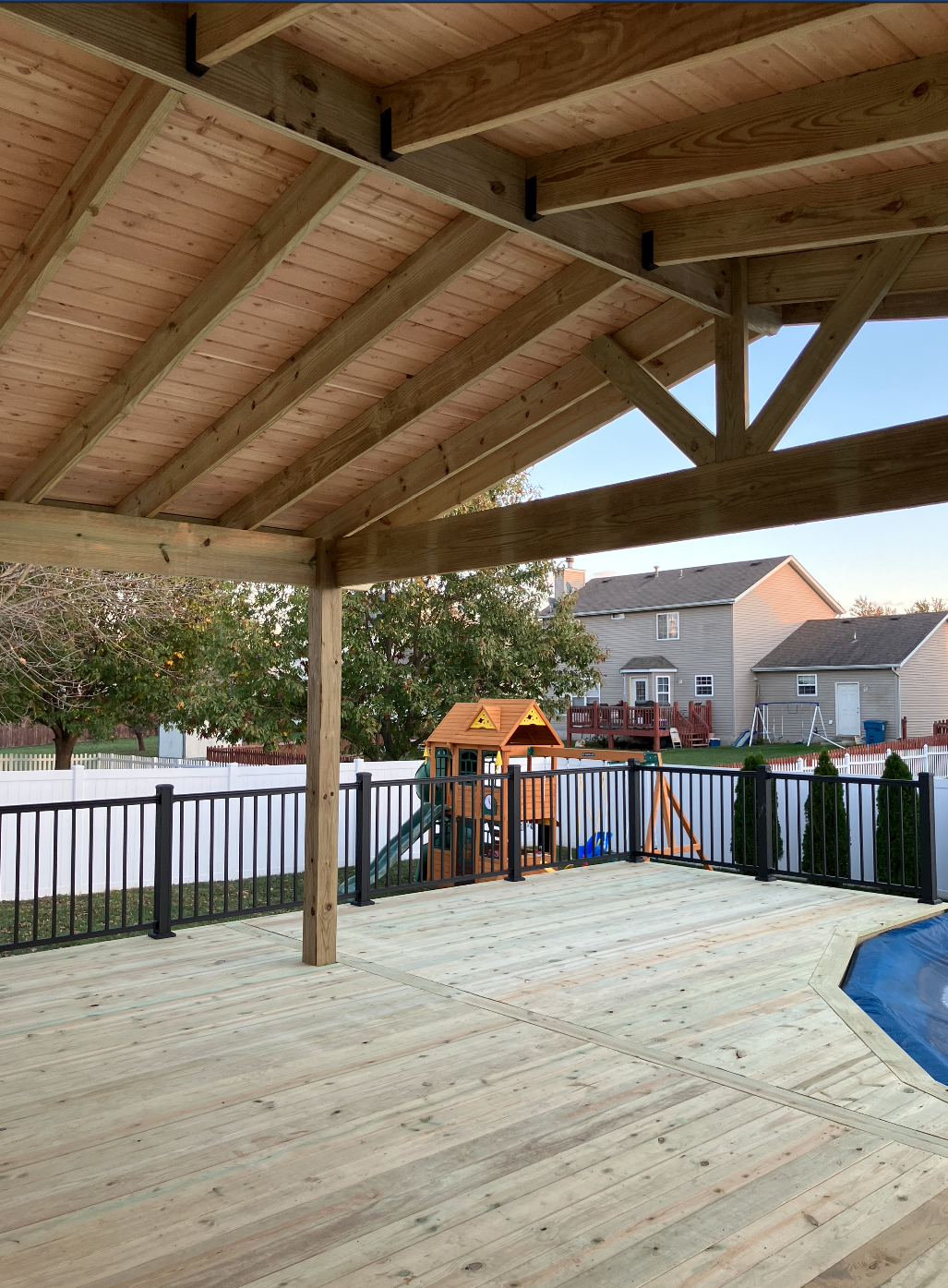 View of a backyard with a newly built wooden deck, metal railing, children's playhouse, swing set, trees, and neighboring houses under a clear sky.