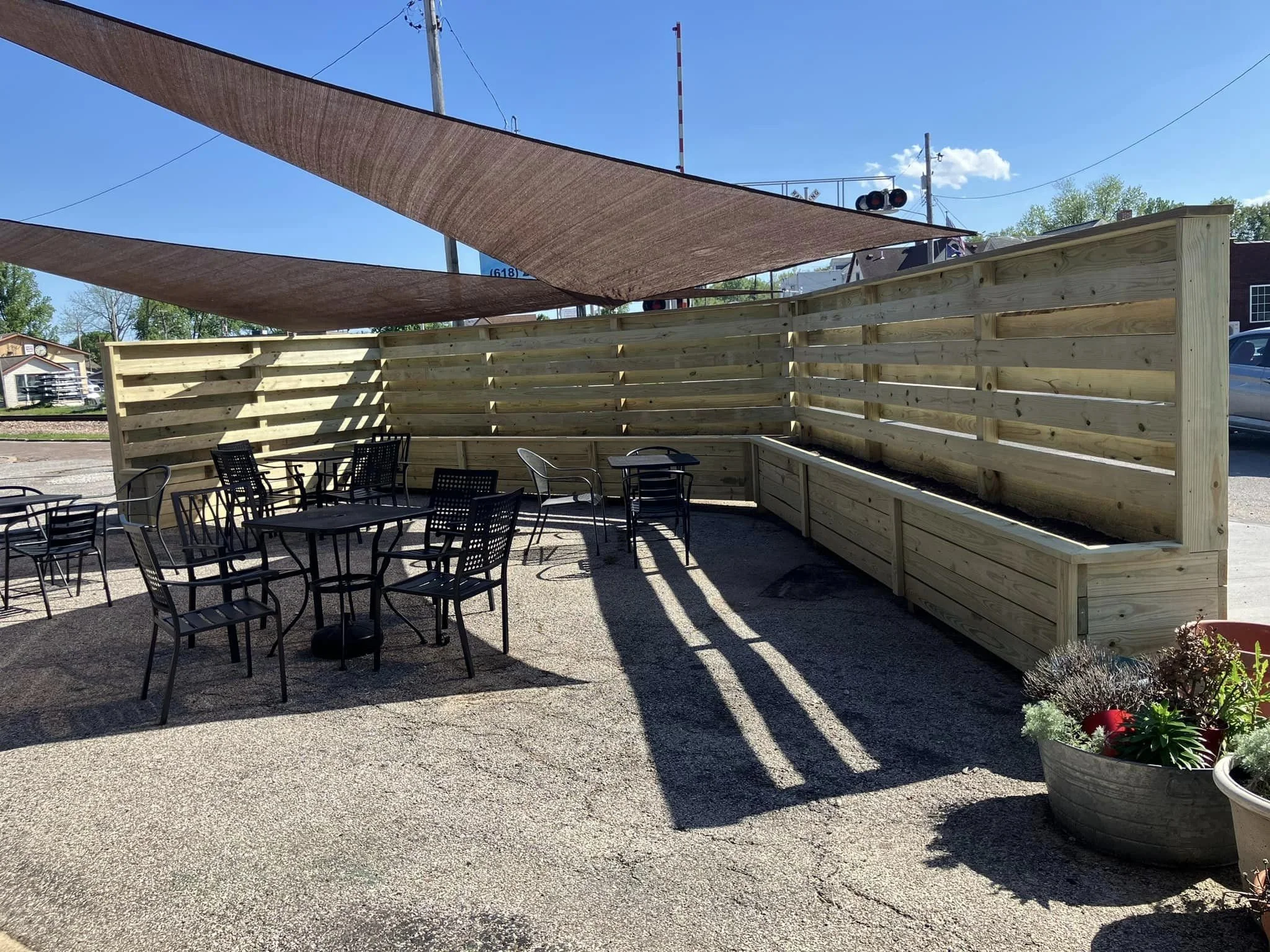An outdoor patio area with black metal chairs and tables, a wooden privacy fence, and sunshade sails overhead. There is a large planter with flowers in the foreground and trees and buildings in the background.