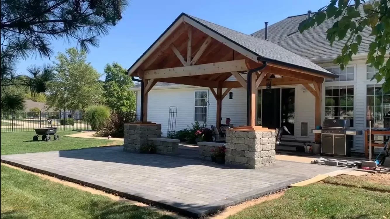 Backyard patio with a covered pergola, stone pillars, outdoor grill, and surrounding lawn and trees.
