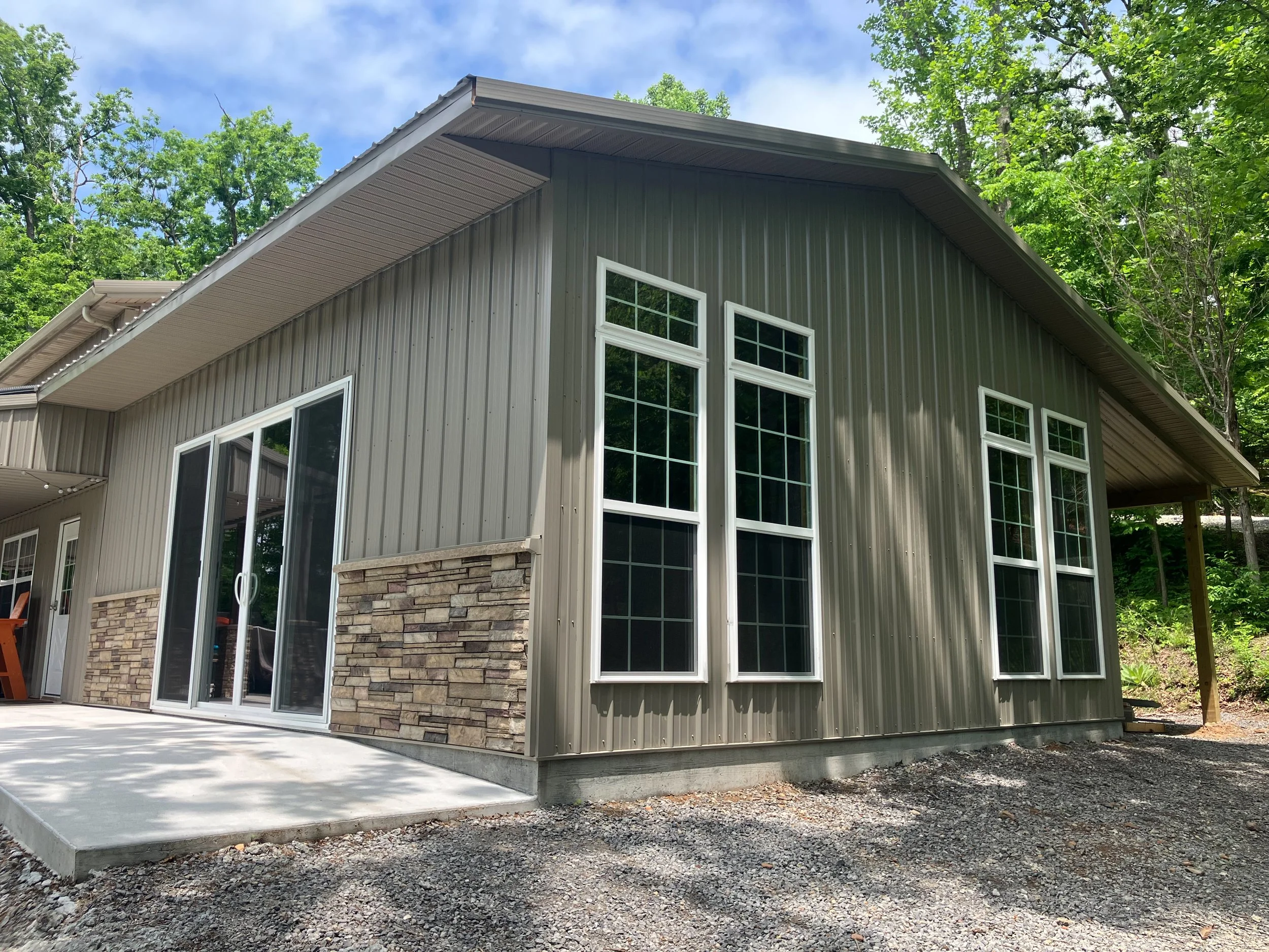 A residential house with metal siding and large windows, surrounded by green trees and a gravel yard, under a partly cloudy sky.