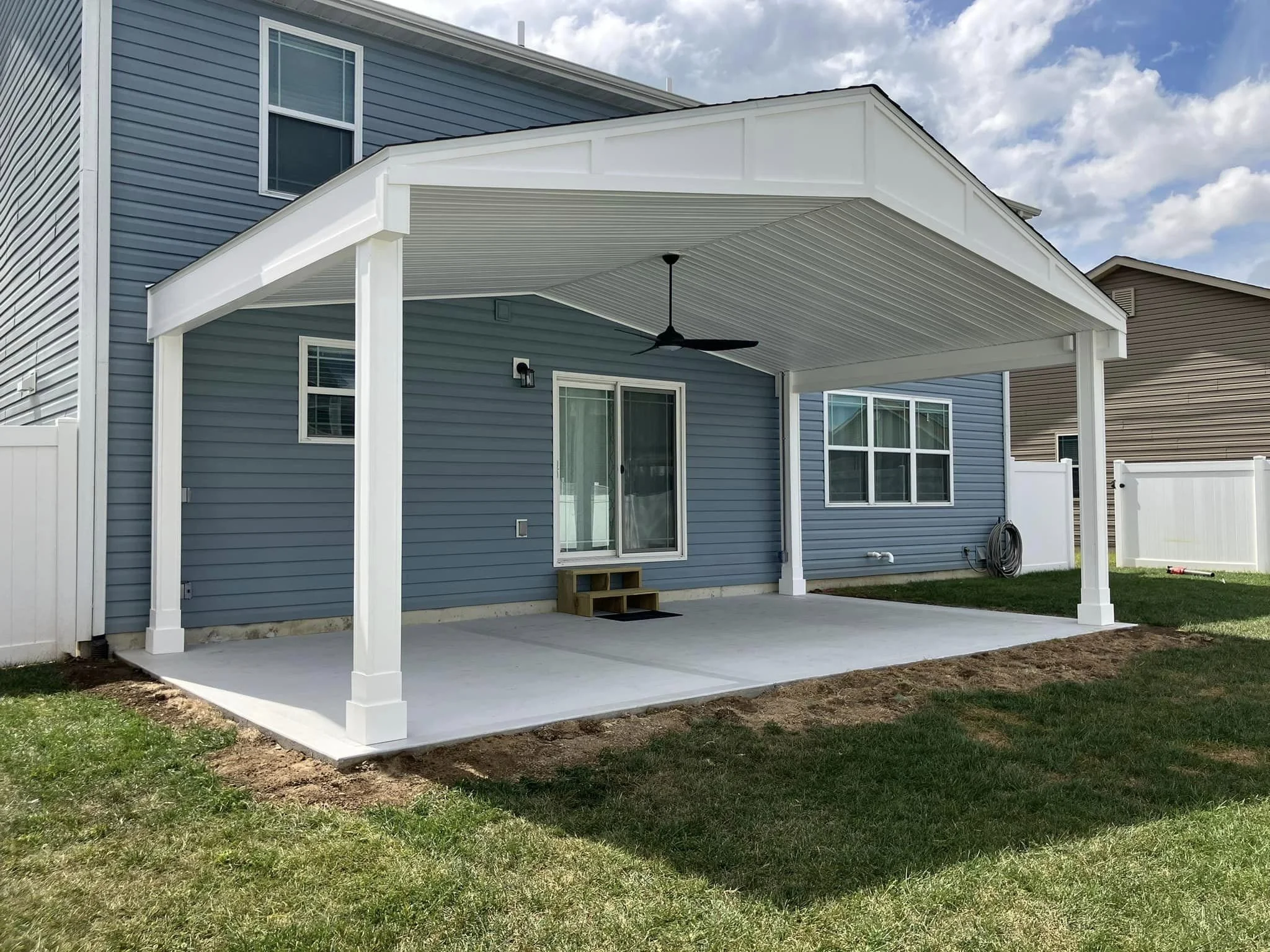 Backyard patio with a white pergola, sliding glass door, and ceiling fan, adjacent to a blue house with multiple windows, a white fence, and a green lawn.