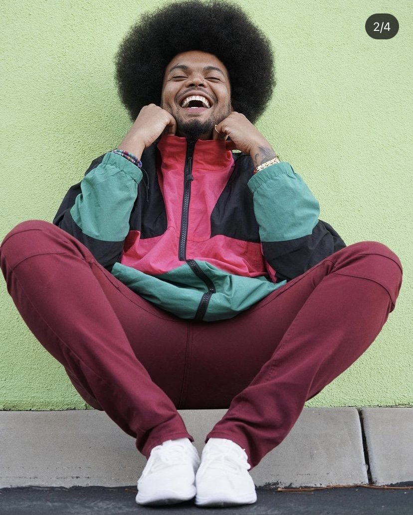A young man with an afro hairstyle sitting on the ground, smiling and adjusting his colorful windbreaker jacket with a green and red background.