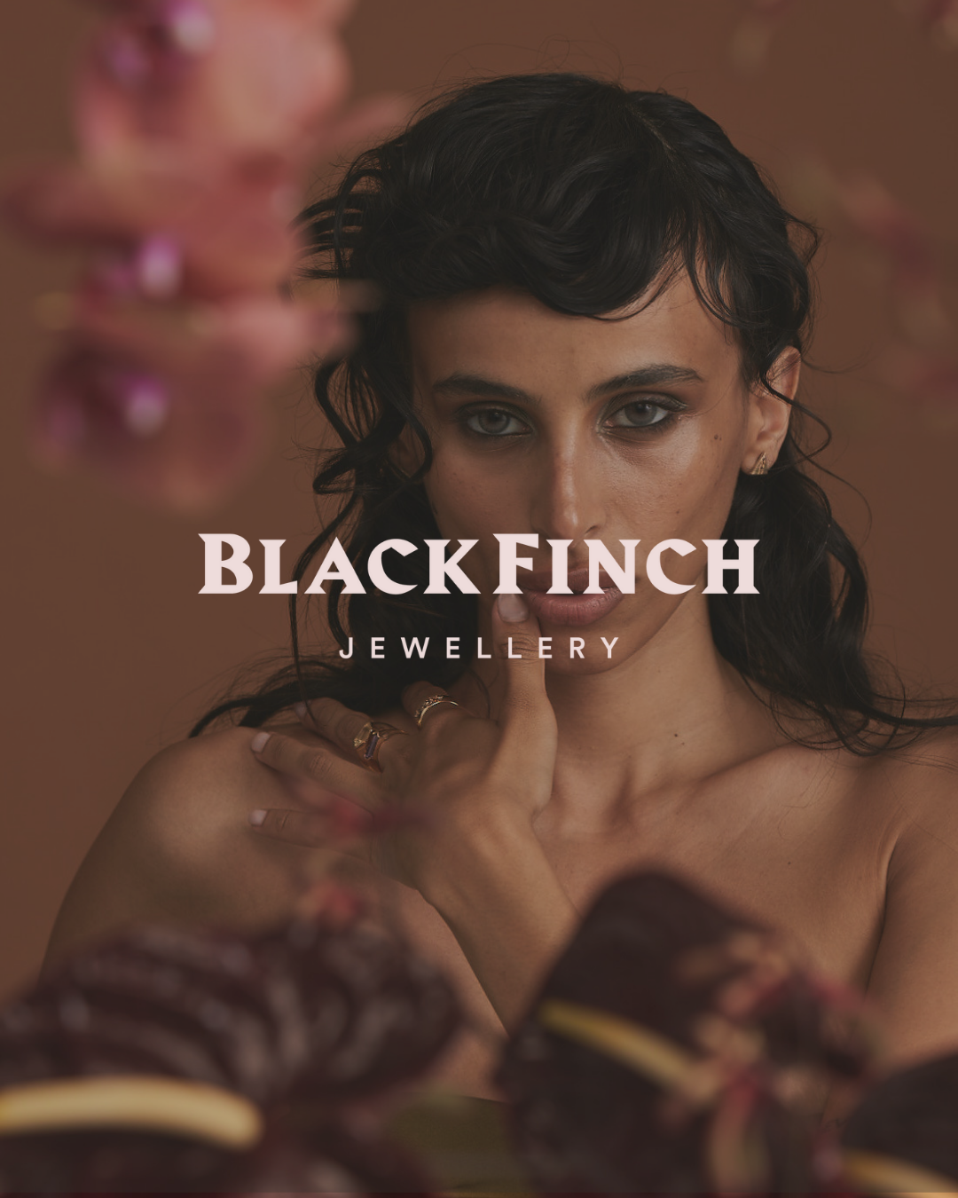 Model posing with Black Finch Jewellery logo, wearing rings, with blurred pink flowers in the foreground.