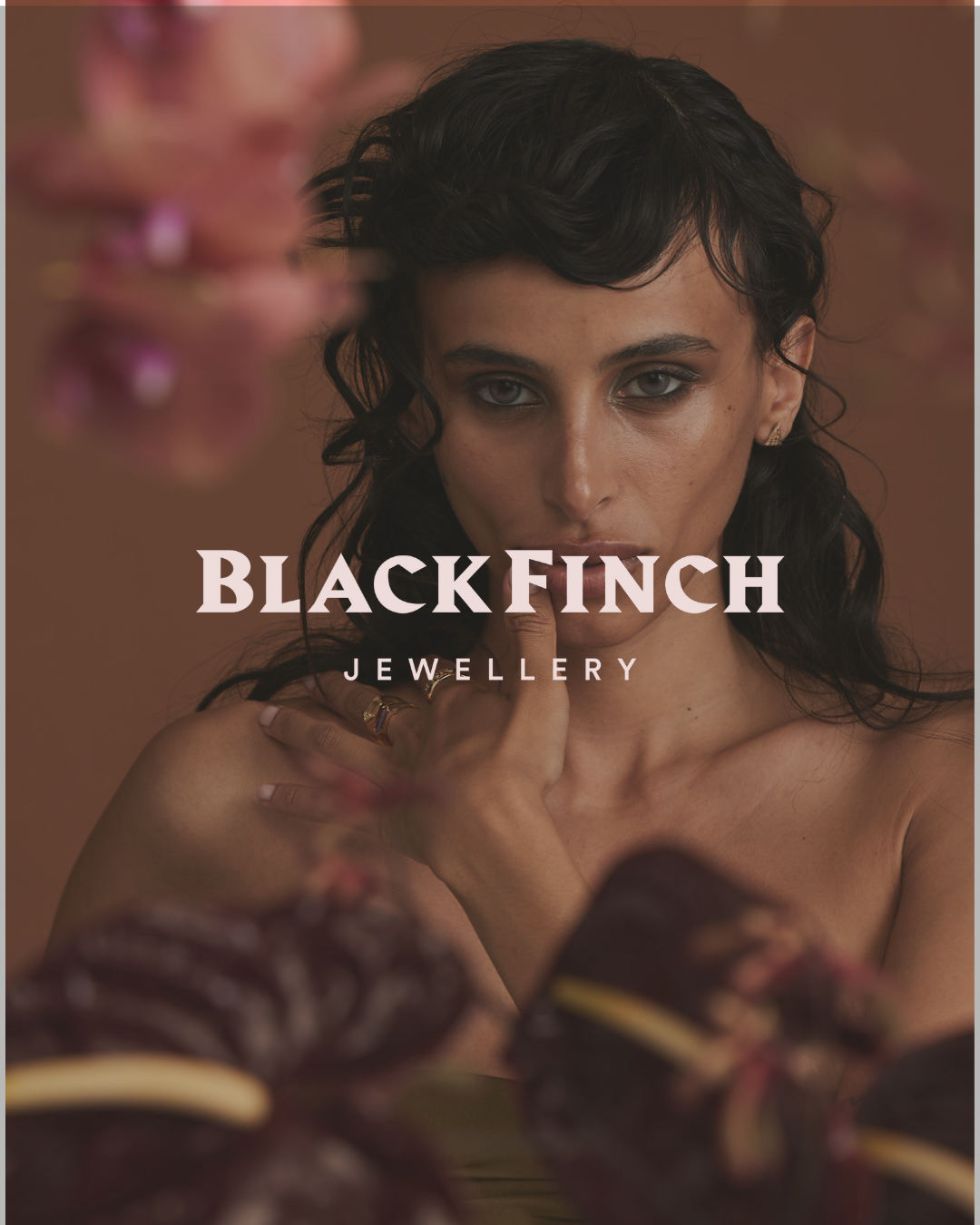 Model posing with Black Finch Jewellery logo, wearing rings, with blurred pink flowers in the foreground.