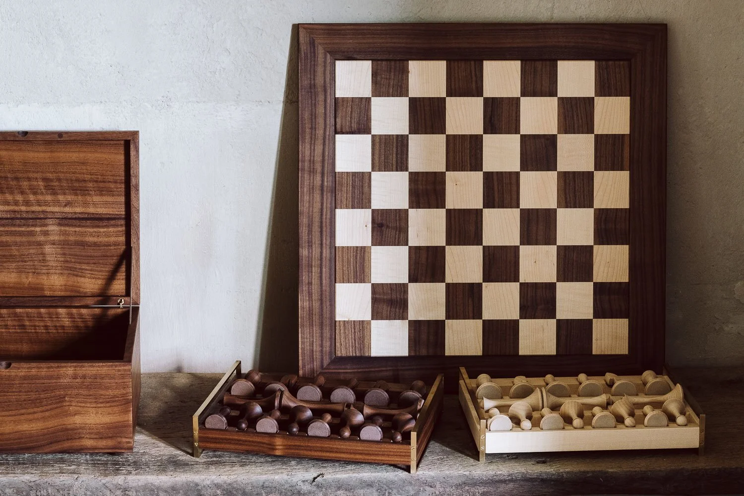 A wooden chessboard with alternating light and dark squares, two trays of chess pieces (dark and light), and a wooden box, all placed on a rustic wooden surface.