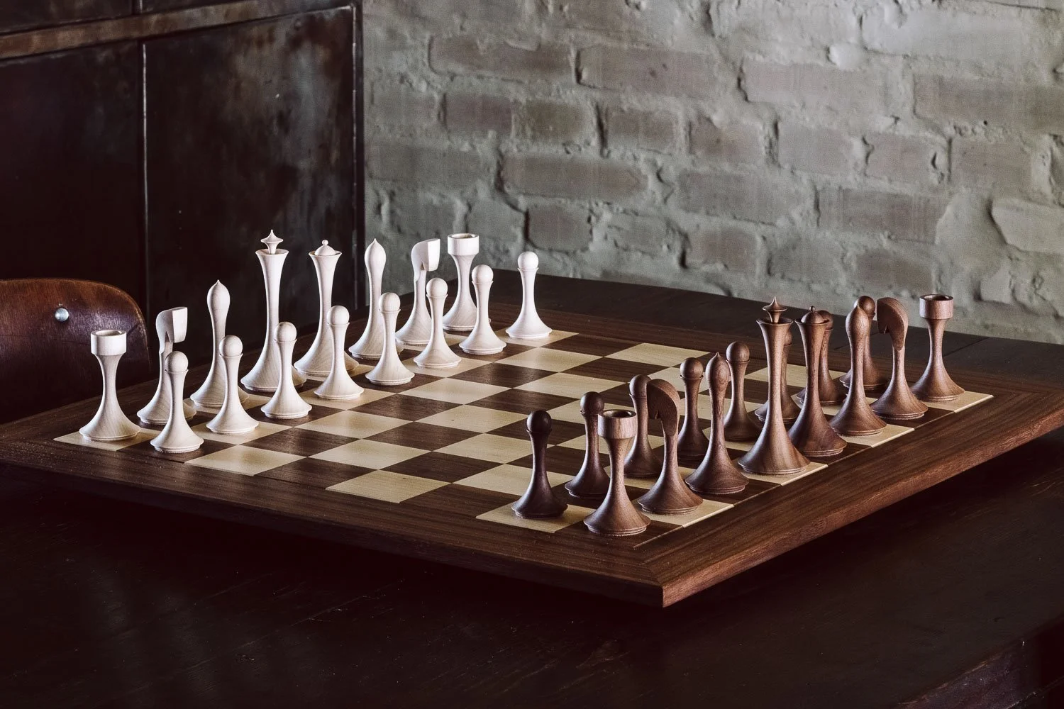 A wooden chessboard with white and brown chess pieces set up for a game, placed on a dark wooden table against a brick wall background.