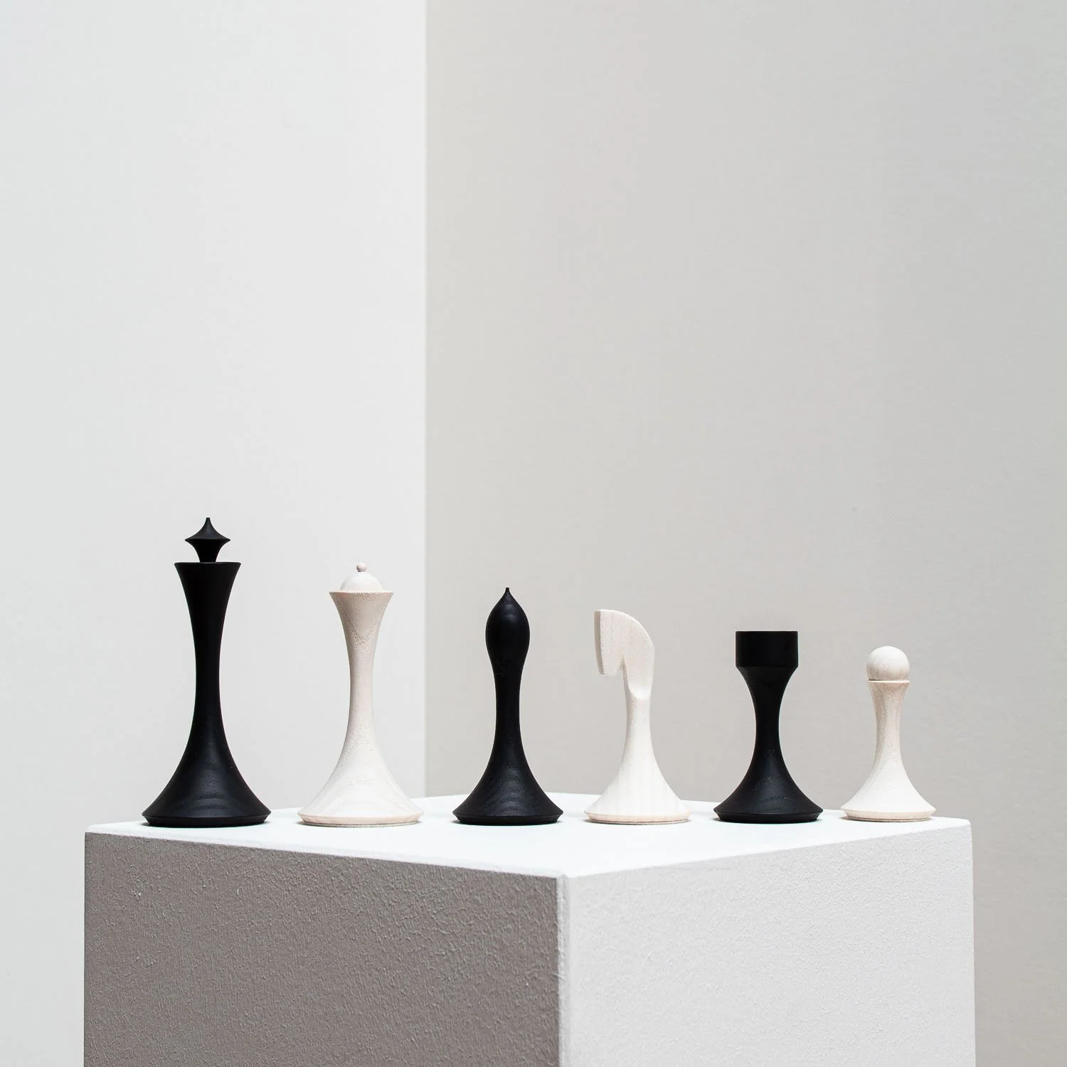 Set of black and white chess pieces on white pedestal against plain wall.