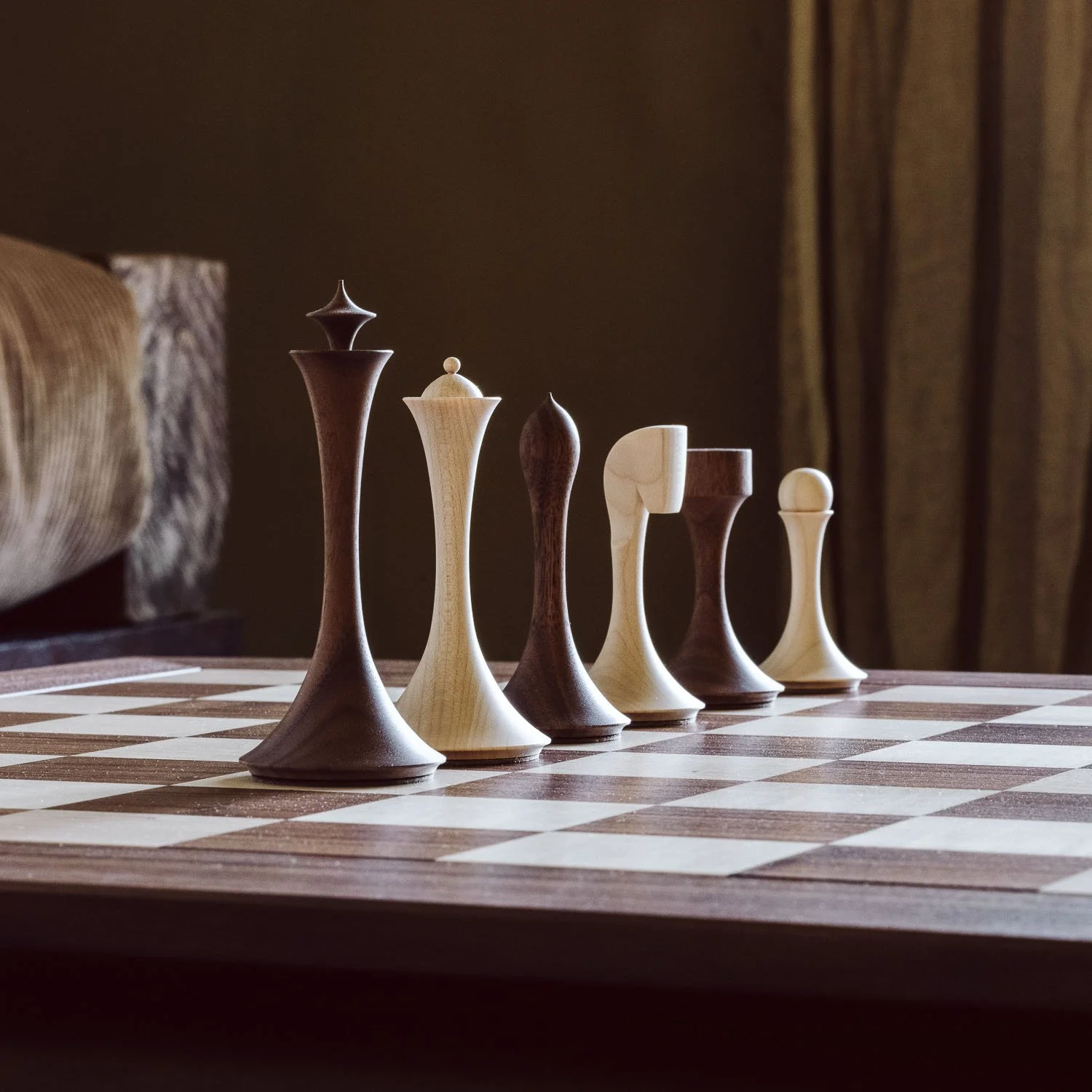 A chessboard with six wooden chess pieces arranged in a row, with a beige curtain and a dark wall in the background.