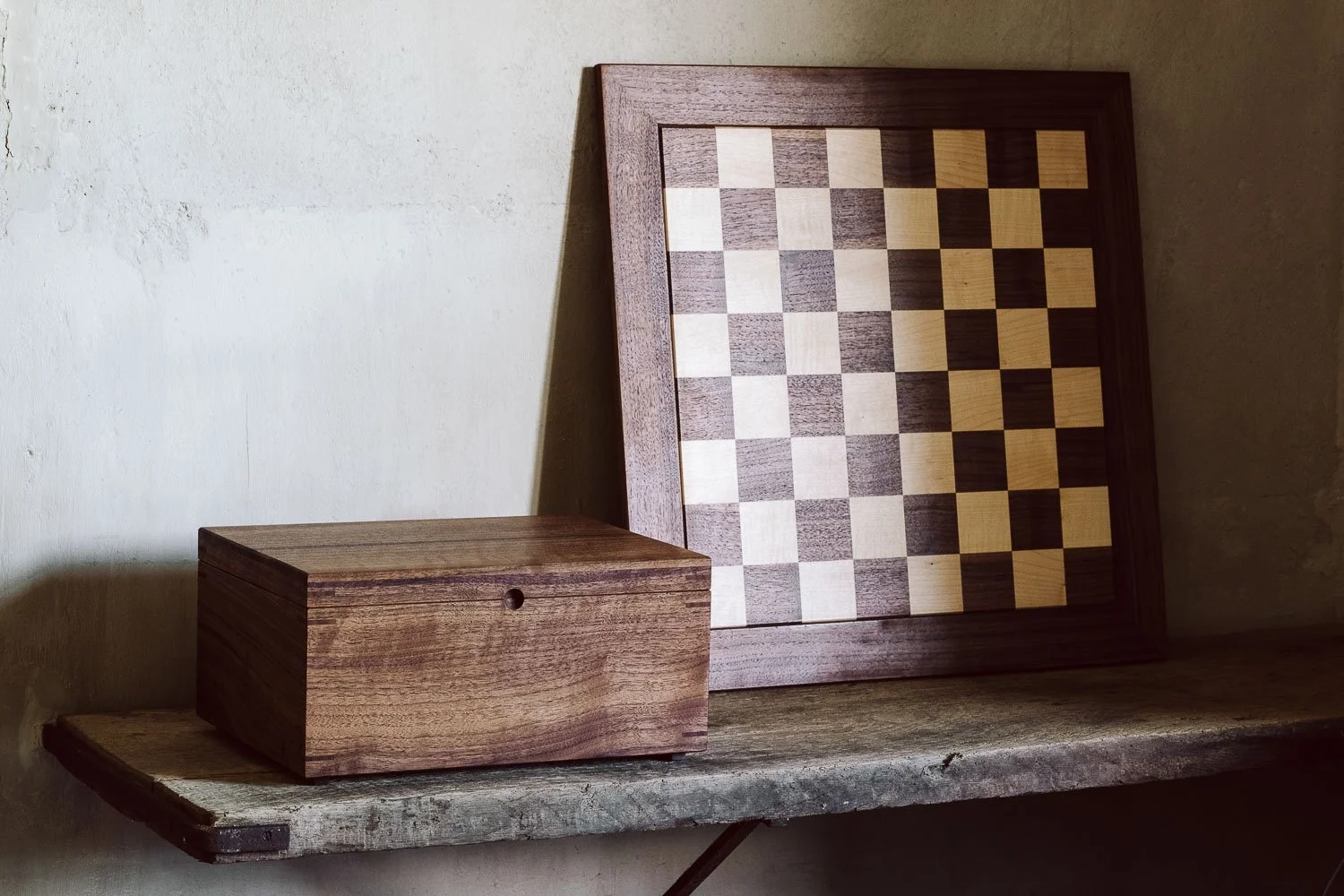 A wooden chessboard with alternating light and dark squares, leaning against a wall on a rustic wooden shelf, with a matching wooden box nearby.