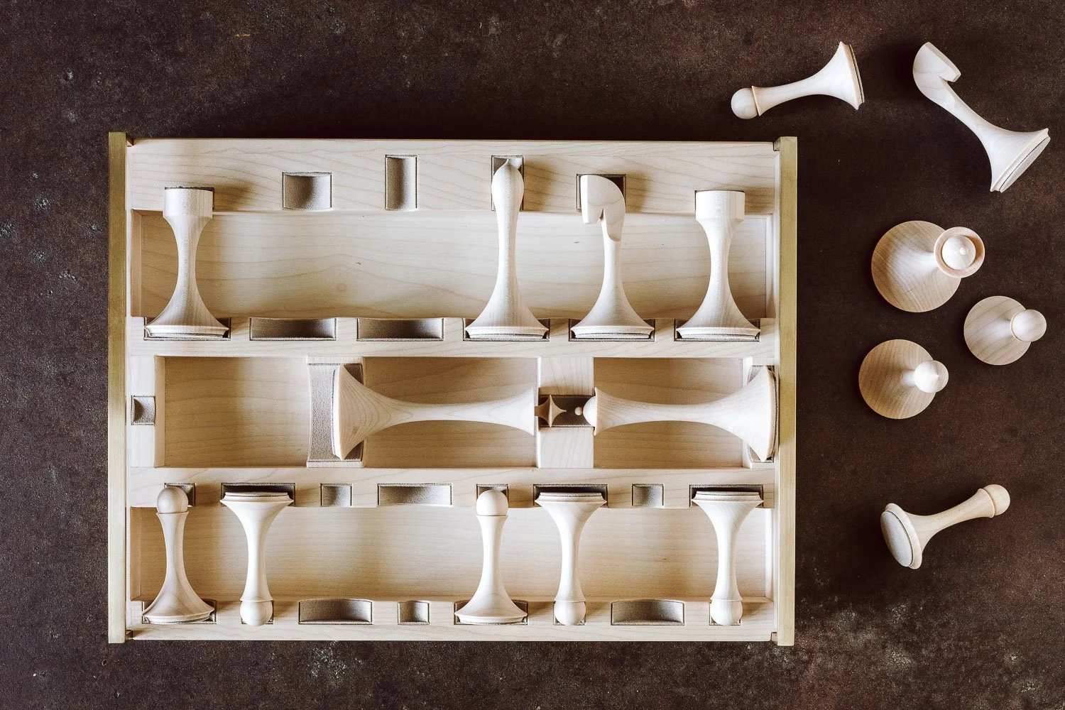 Unfinished wooden chess set with pieces arranged in a wooden storage box, some pieces placed on a dark surface.