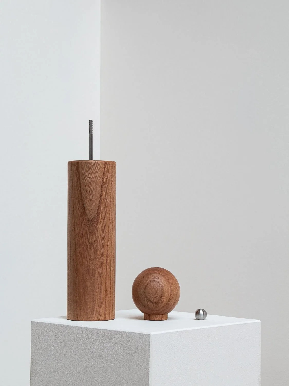 Minimalist art display with a tall wooden cylinder, a small wooden sphere, and a small metal ball on a white pedestal against a plain light gray background.