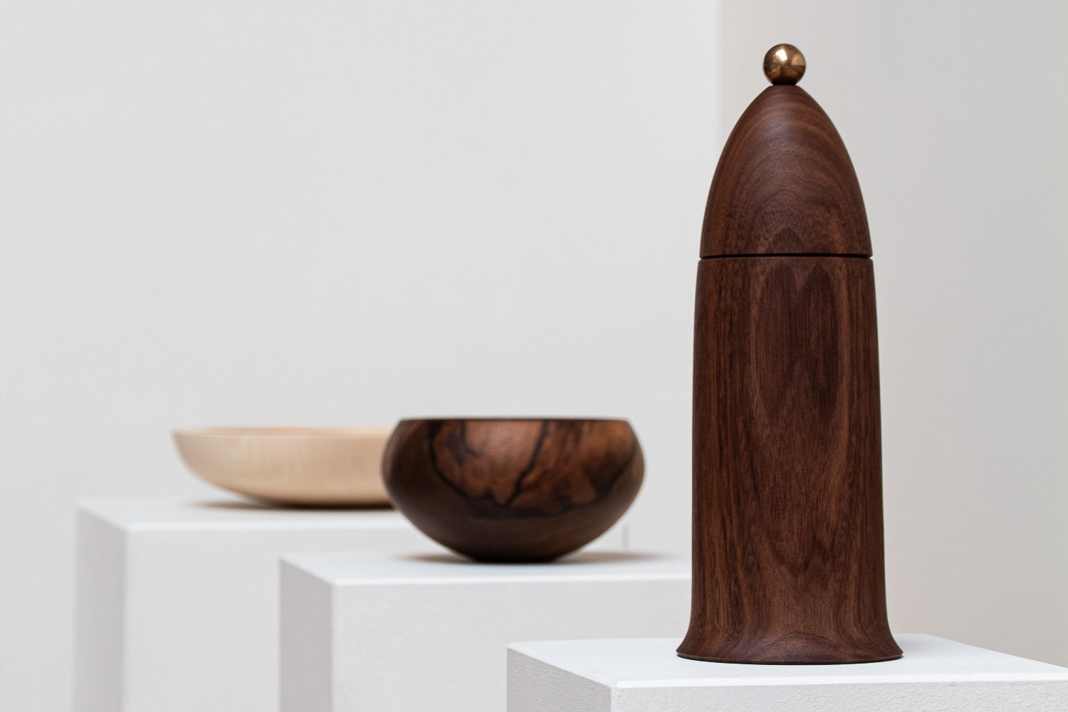 Three wooden art pieces displayed on white pedestals, including a tall cylindrical sculpture with a rounded top and a small sphere, and two bowls in light and dark wood.