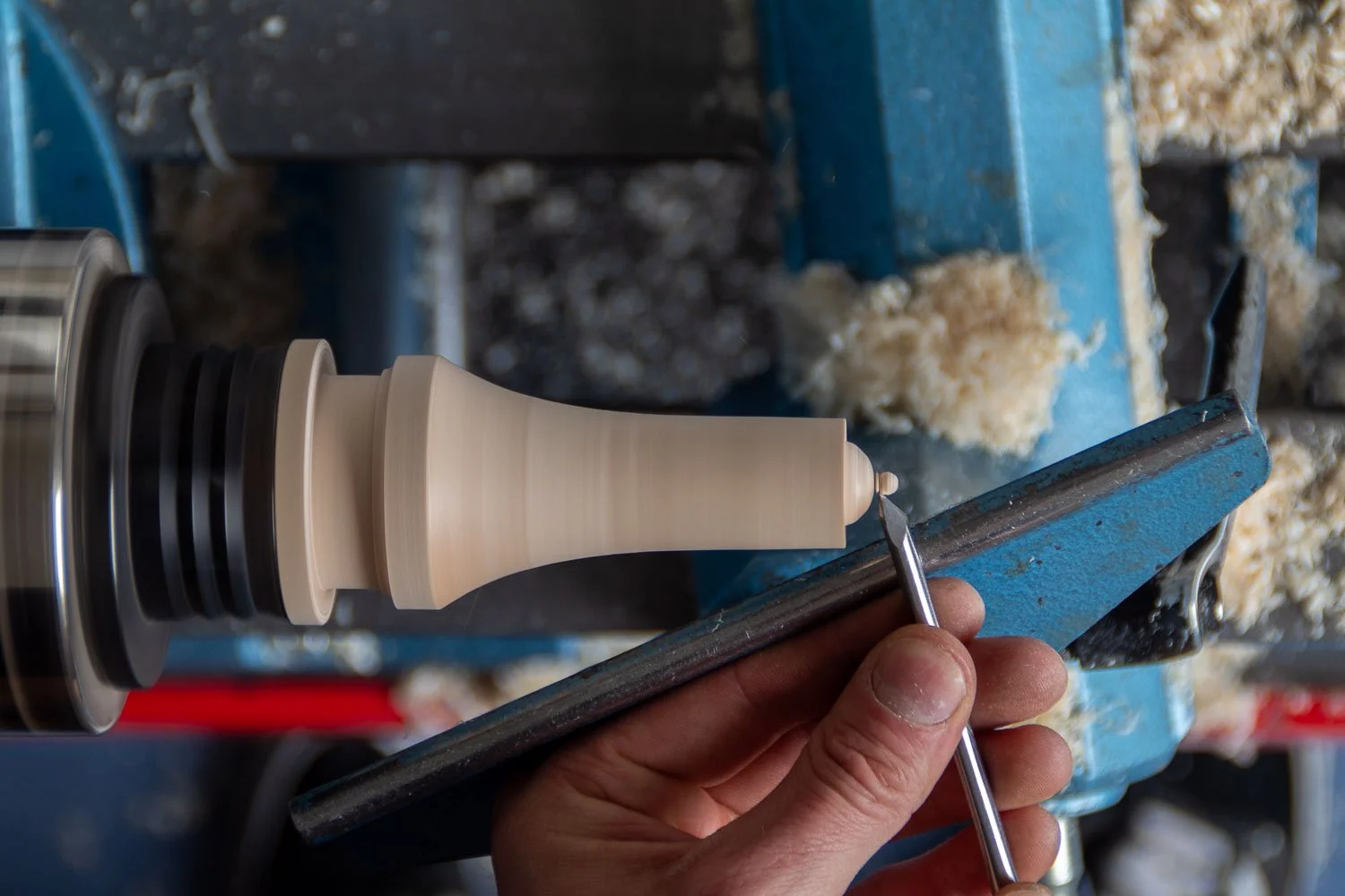 A person is carving a decorative wooden object on a lathe, using a small chisel, with wood shavings around.