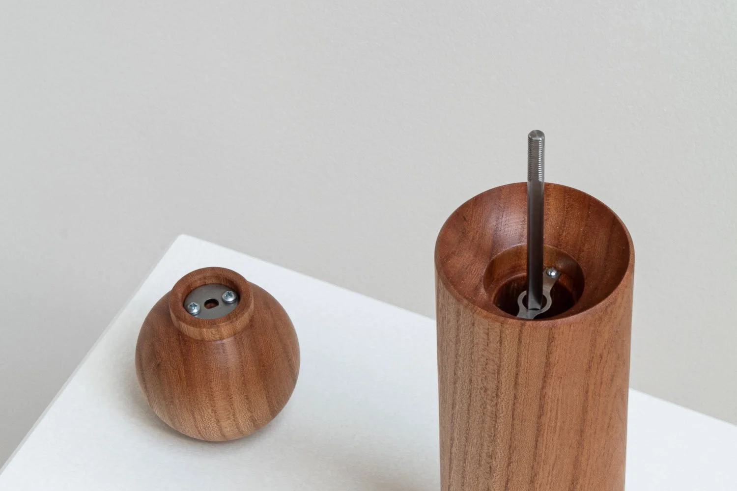 A wooden pepper grinder with its top removed, exposing the grinding mechanism, on a white surface against a plain wall.