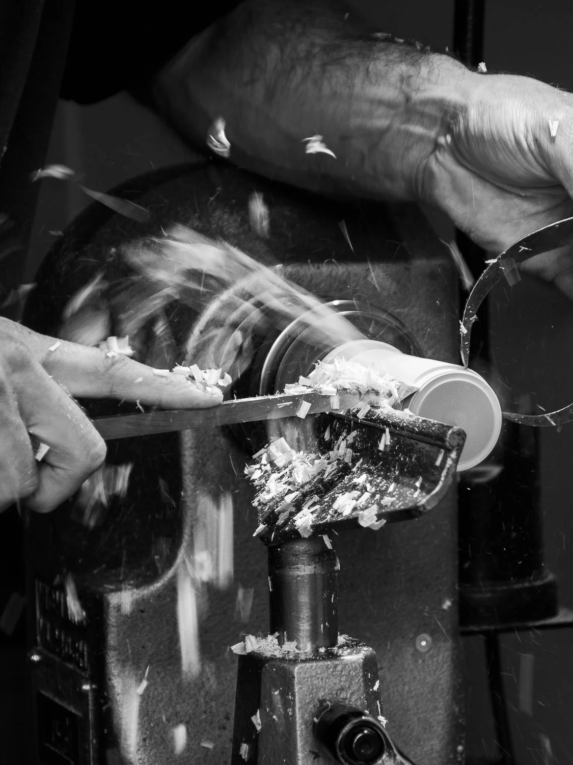 A person is using a hand lathe machine to carve or shape a cylindrical piece of material, with wood shavings flying as the cutter rotates.