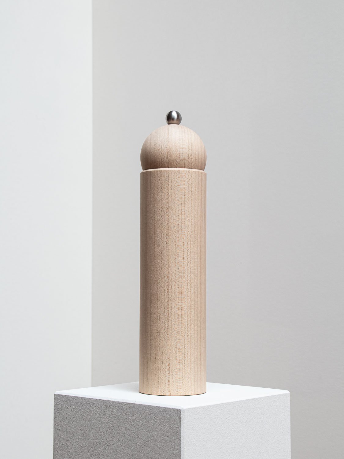 Wooden pepper mill with a metal top on a white pedestal against a plain light-colored background.
